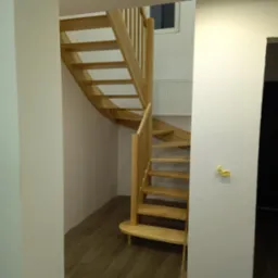 Spiralne schody drewniane z jasnego drewna, prowadzące w górę, widoczne z perspektywy dolnej części schodów. Na ścianie widoczne ślady po pracach montażowych.