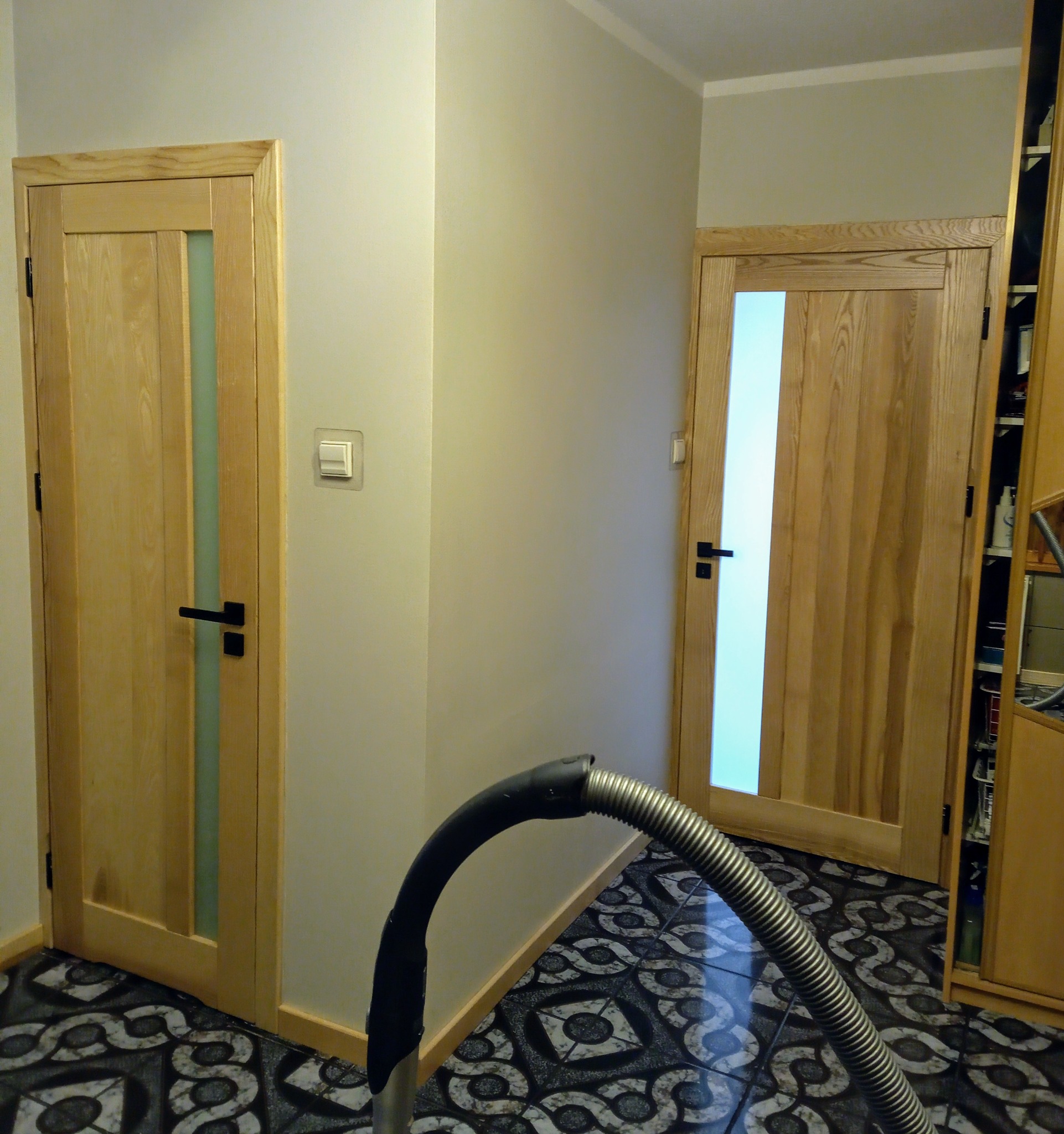 Dwa jasne, drewniane drzwi z przeszkleniami wstawione w jasnym wnętrzu. Na podłodze wzorzysta posadzka. Widoczny fragment odkurzacza.