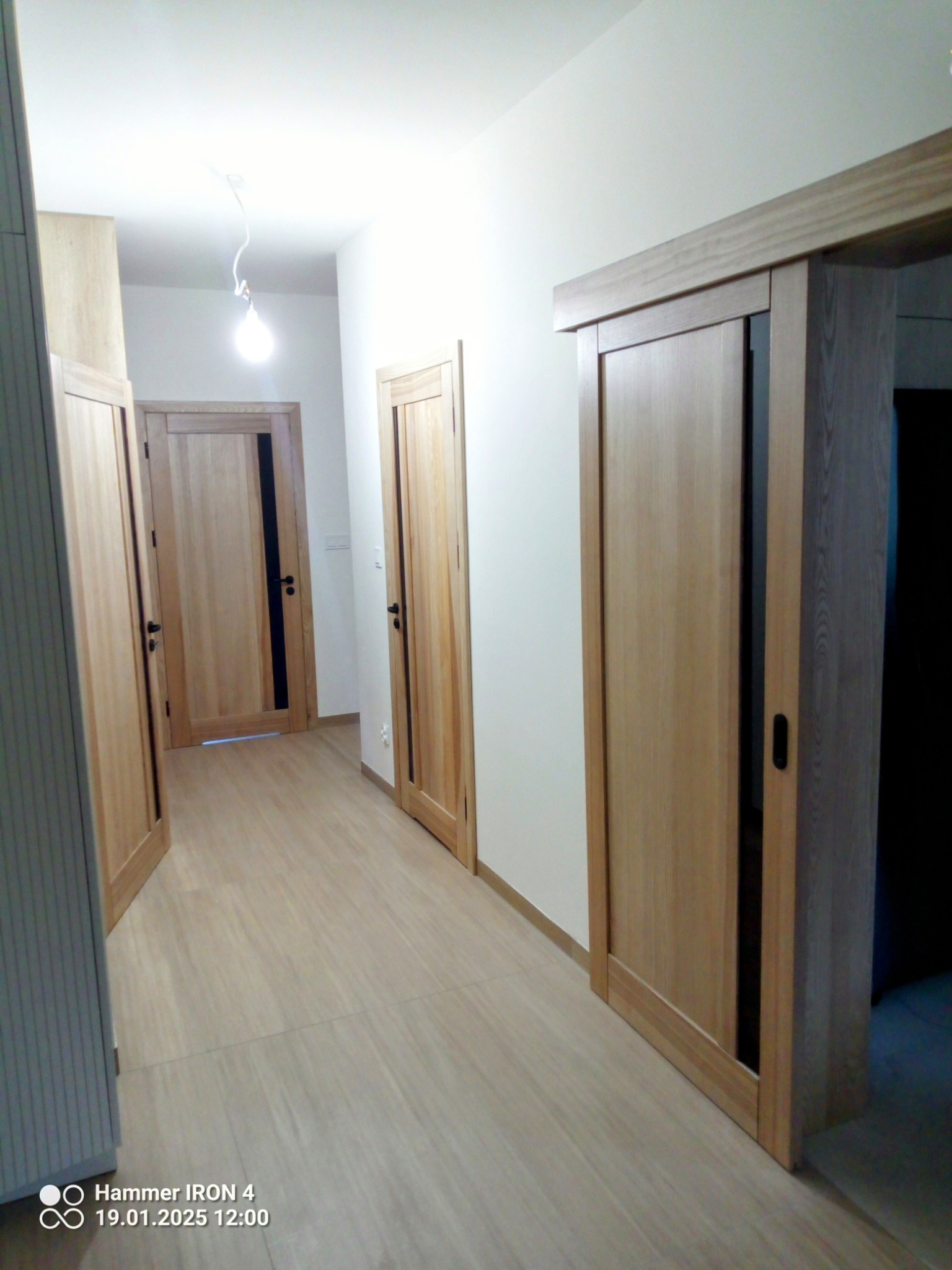 Jasne, drewniane drzwi przesuwne z czarnym uchwytem, widoczne w korytarzu z trzema parami drzwi po lewej stronie, na podłodze jasne płytki imitujące drewno.