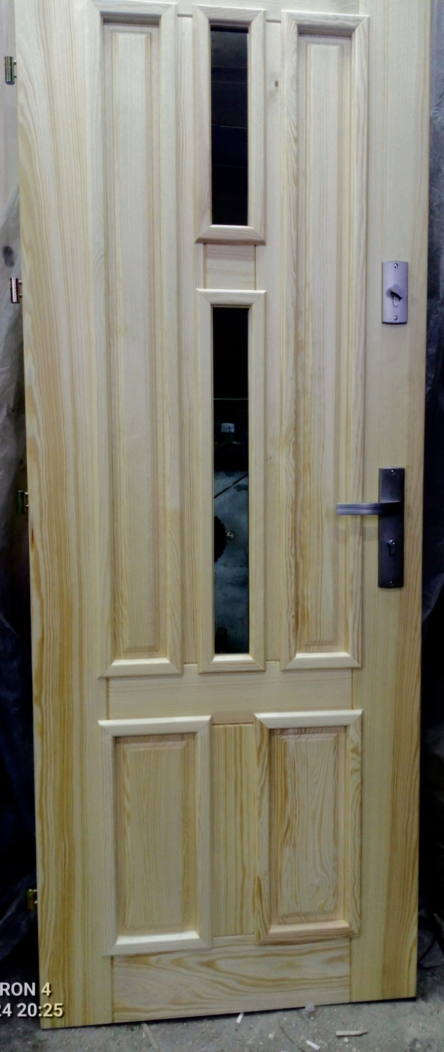 Drewniane drzwi wejściowe z szybkami, surowe, bez wykończenia. Widoczne okucia i zamek. Charakterystyczny wzór drewna.