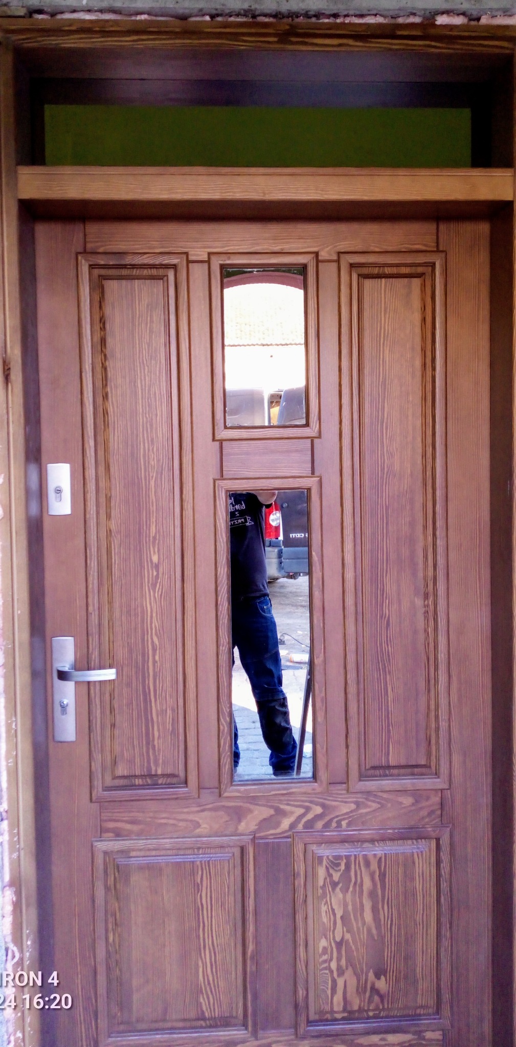 Frontowe, brązowe drzwi wejściowe z szybkami, widoczny srebrny zamek i klamka. W szybach odbija się sylwetka osoby i fragment otoczenia.