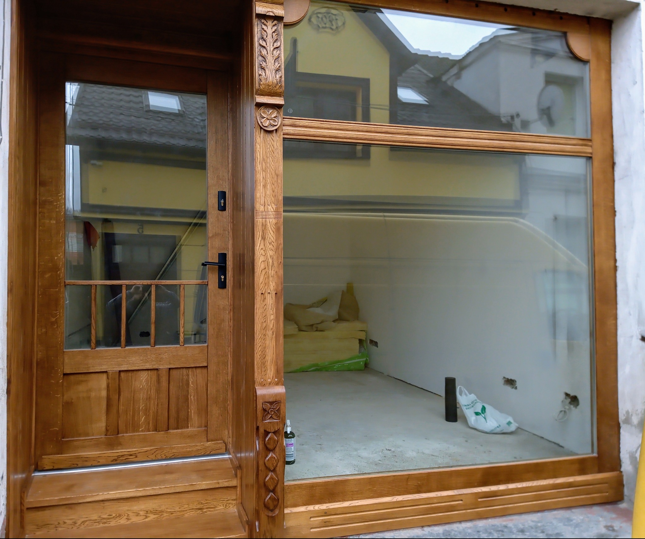 Otwarta, przeszklona witryna z dębową ramą i zdobieniami, widok na wnętrze w trakcie prac wykończeniowych. W tle budynek mieszkalny.