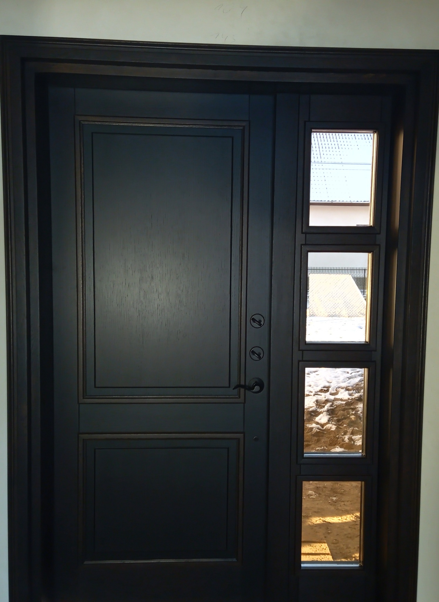 Ciemne, eleganckie drzwi wejściowe z bocznym panelem z trzema okienkami, przez które widać otoczenie na zewnątrz. Klasyczny design z nowoczesnymi akcentami.