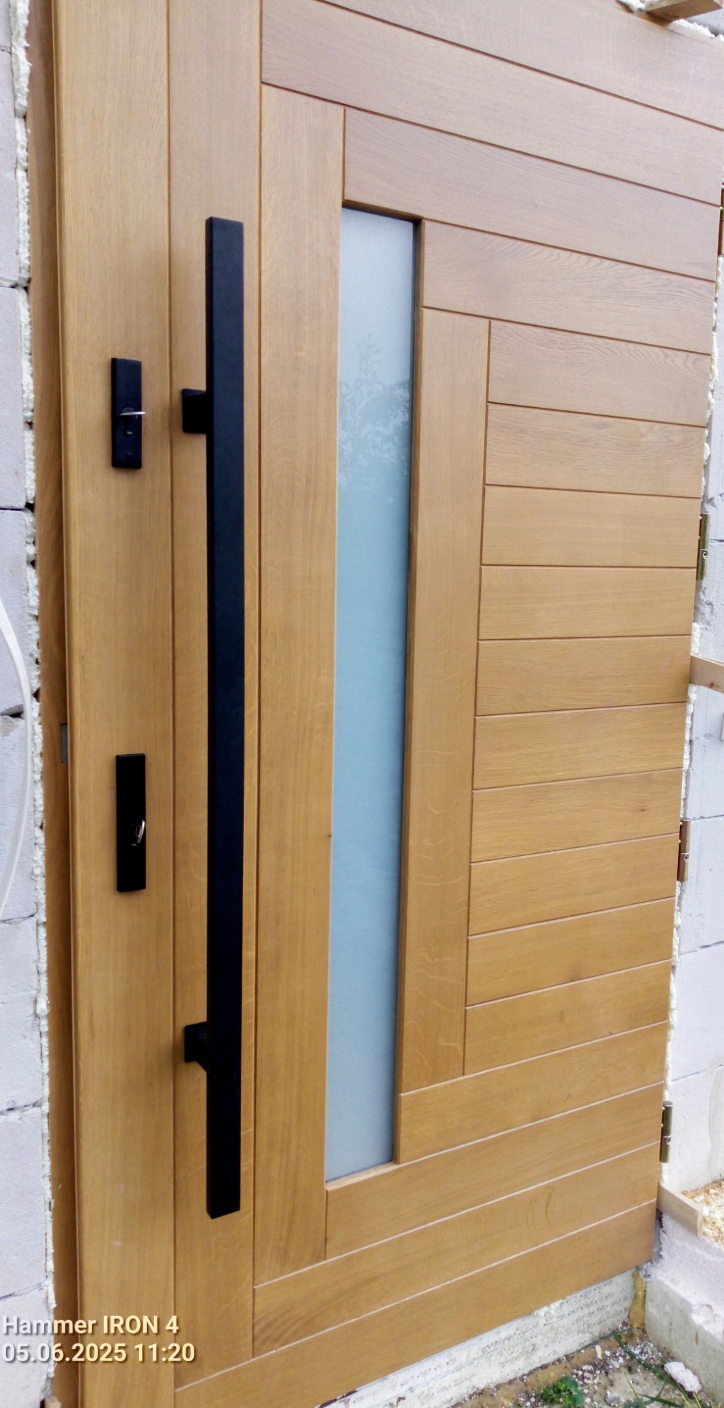 Nowoczesne drzwi wejściowe z drewna w odcieniach brązu, z długim, czarnym uchwytem i pionowym, matowym przeszkleniem, zamontowane w surowym murze.