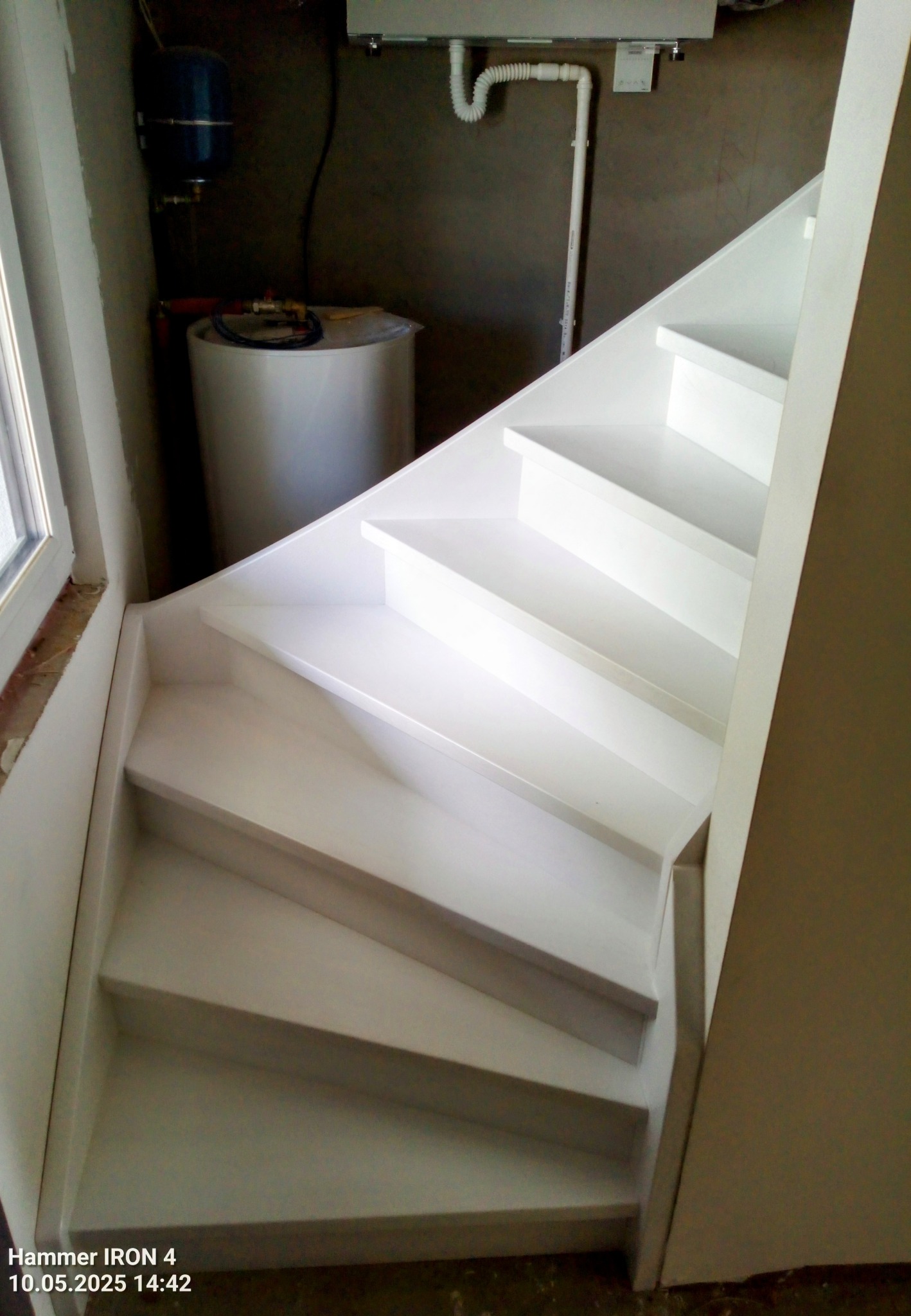 Białe schody prowadzące w dół, z widocznym bojlerem w tle. Ujęcie z dołu, ukazujące zakręt schodów i fragment okna po lewej stronie.