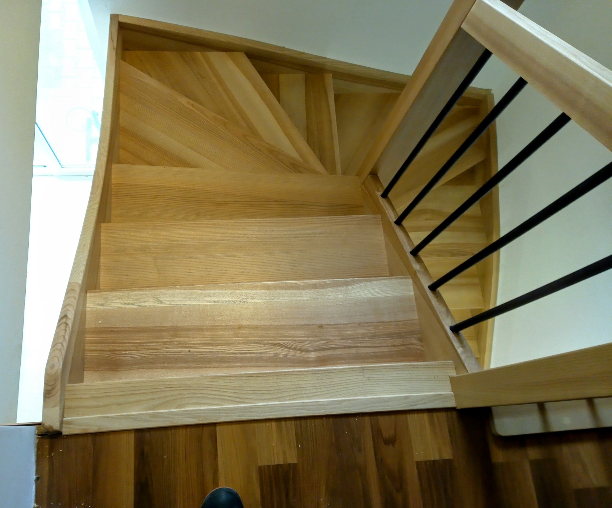 Jasne, drewniane schody z czarnymi balustradami, widziane z góry. Naturalne oświetlenie podkreśla strukturę drewna. Parkiet na dole.