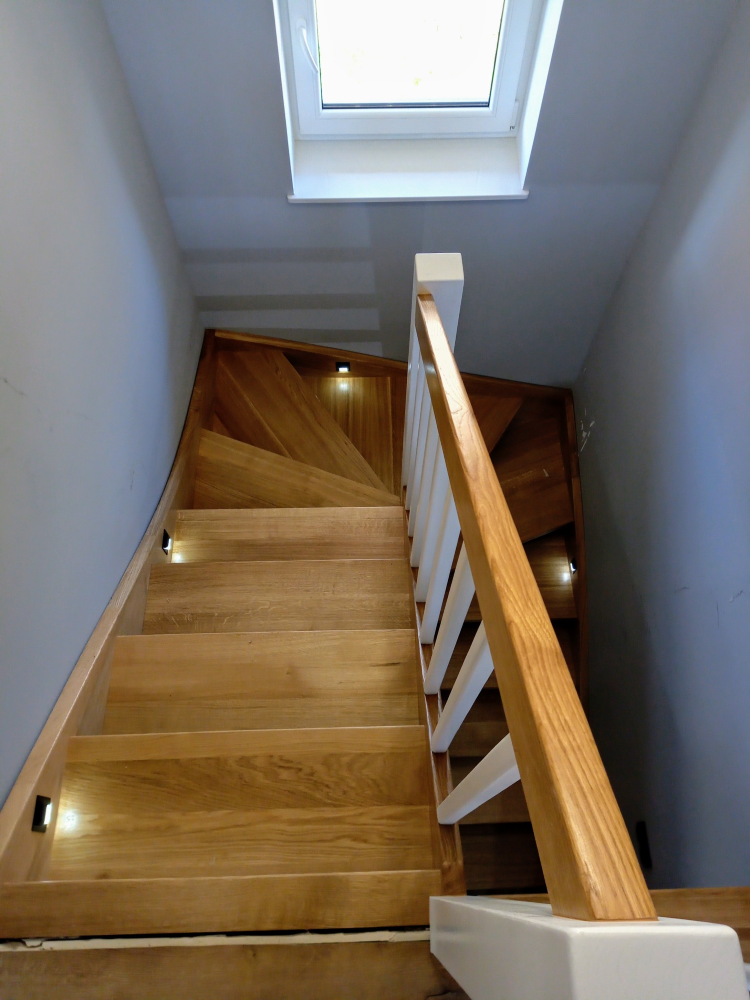 Nowoczesne schody drewniane z oświetleniem LED i białymi balustradami, widok od dołu, oświetlone światłem dziennym z okna dachowego.