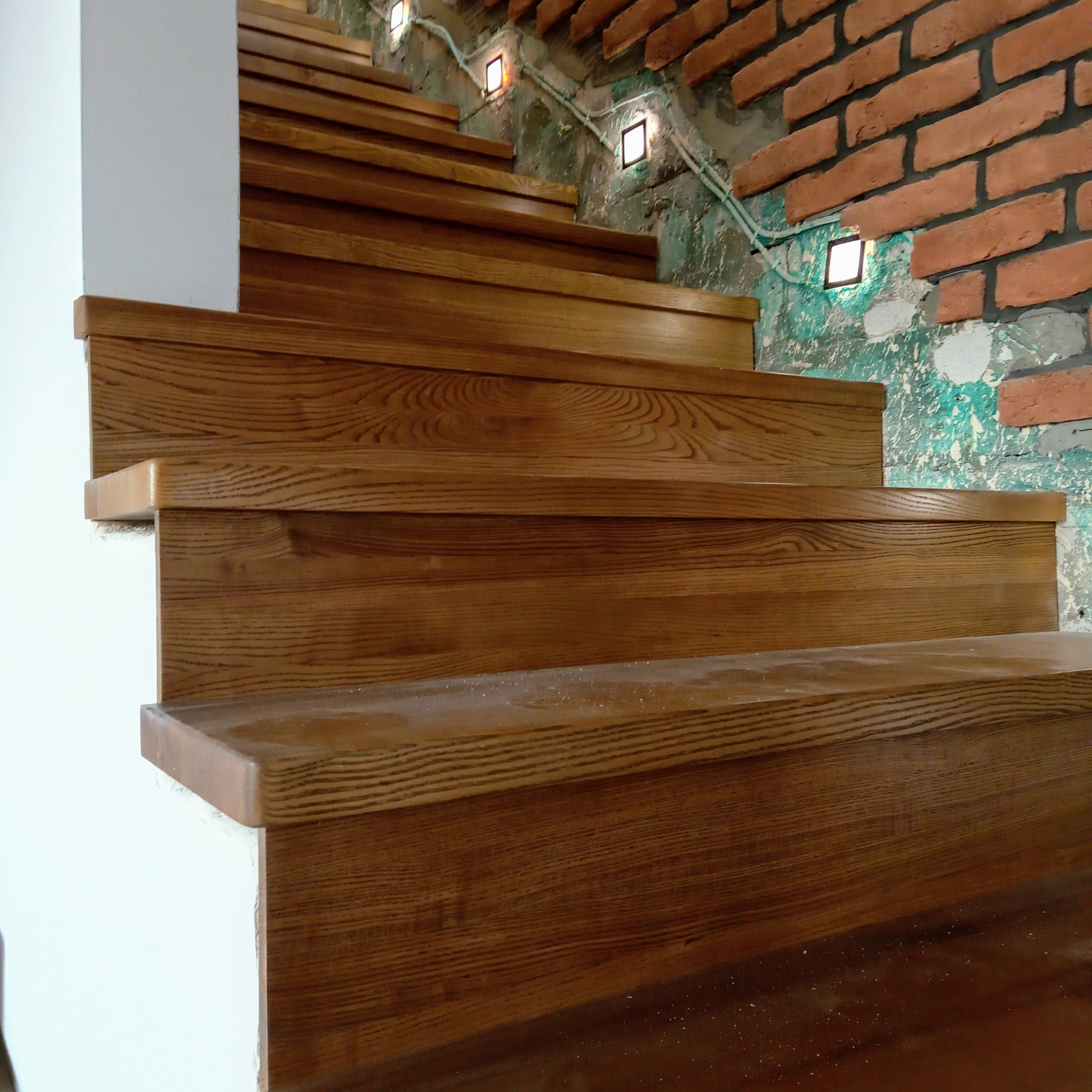 Detal dębowych schodów z oświetleniem punktowym na ceglanej ścianie. Widoczne usłojenie drewna i fragment białej ściany. Ujęcie z dołu.