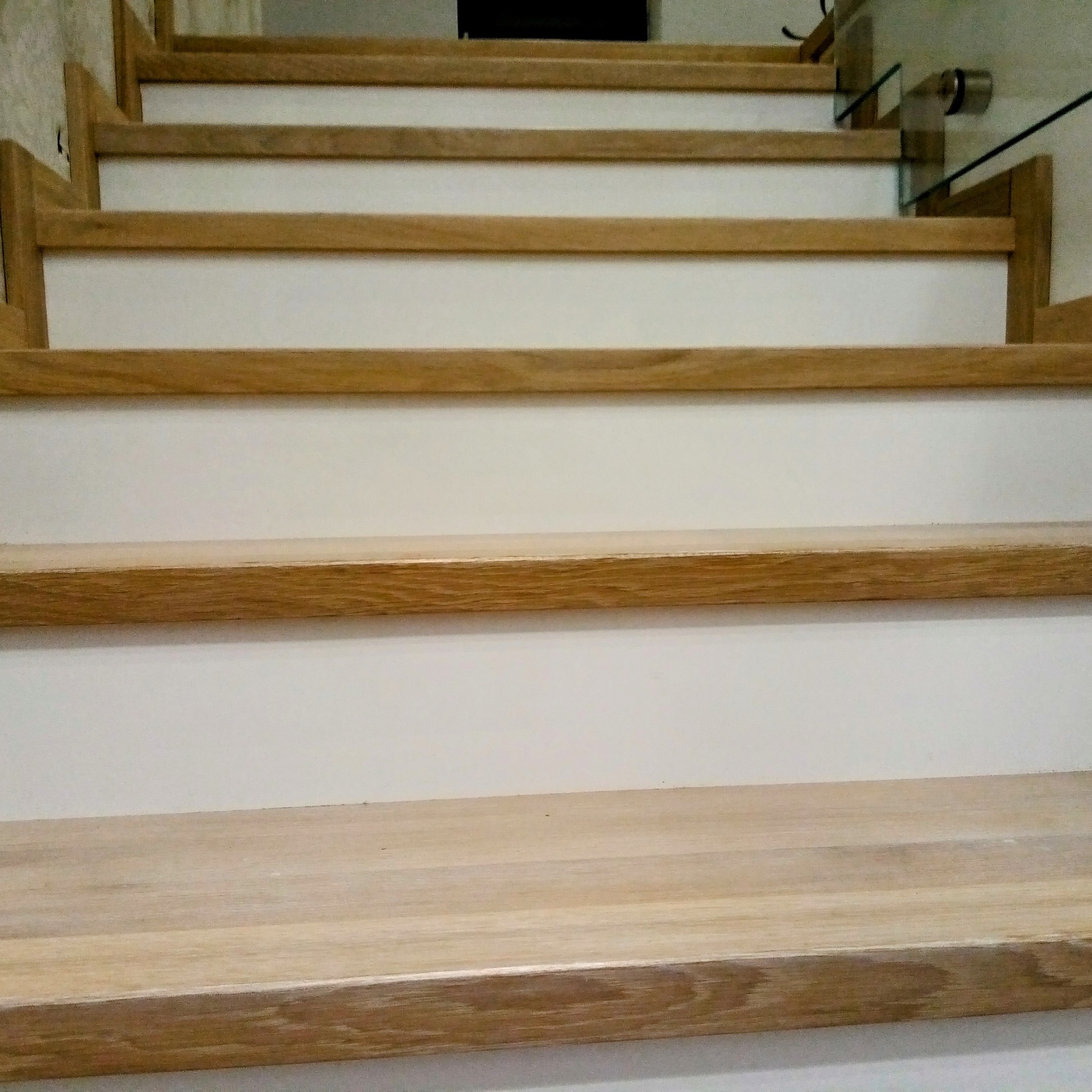 Jasne schody z drewnianymi stopniami i białymi podstopnicami, z widocznymi szklanymi elementami balustrady. Ujęcie z dołu.