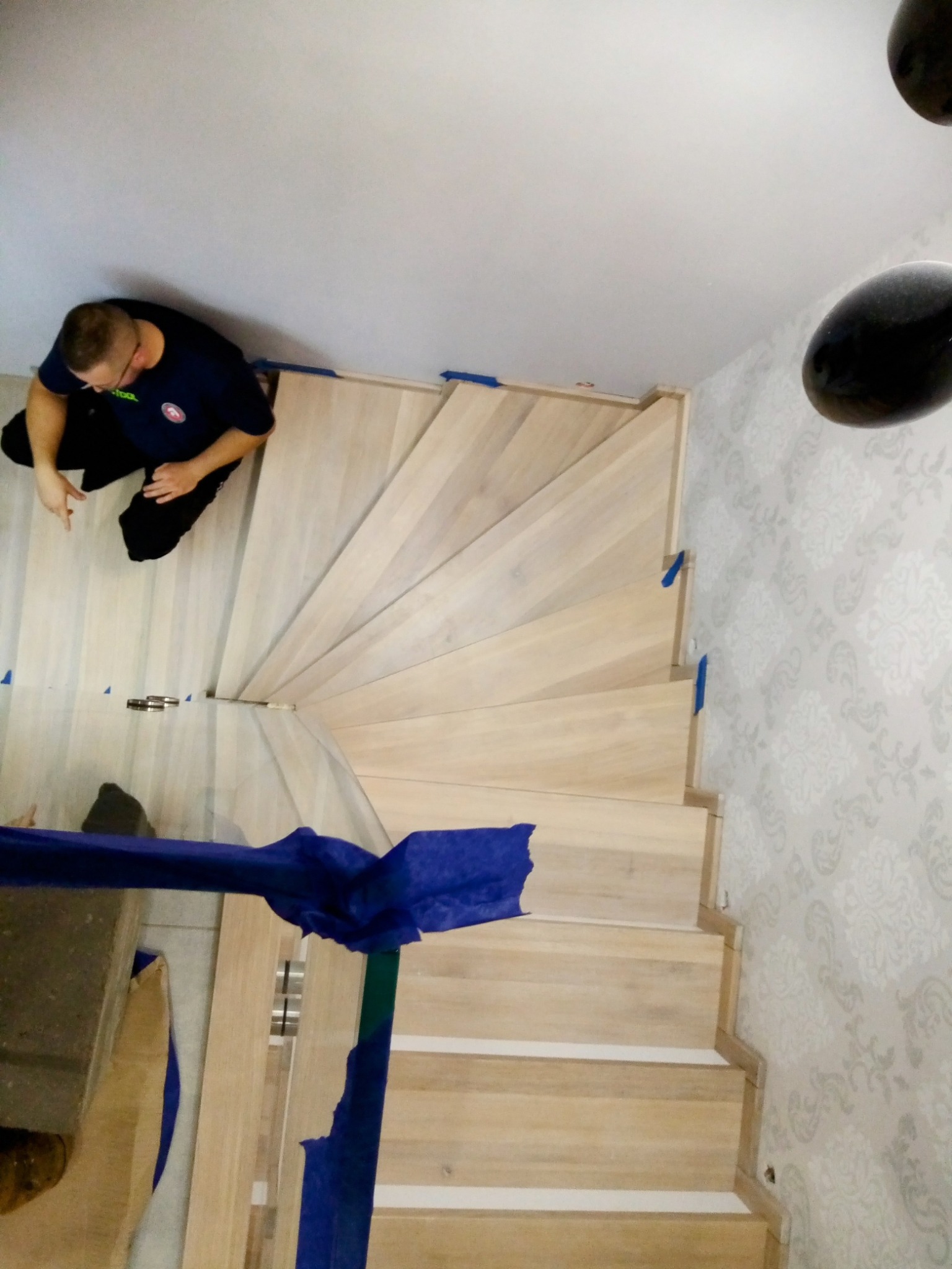 Spiralne schody w trakcie montażu. Widoczny stolarz przy pracy, narzędzia oraz zabezpieczenia na ścianie. Jasne drewno i wzorzysta tapeta w tle.