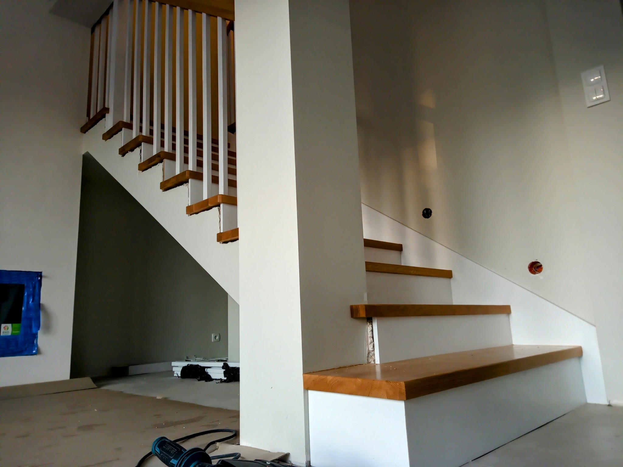 Białe schody z drewnianymi stopniami w nowoczesnym wnętrzu. Widoczne elementy montażowe, podkreślające etap prac wykończeniowych. Minimalistyczny design.