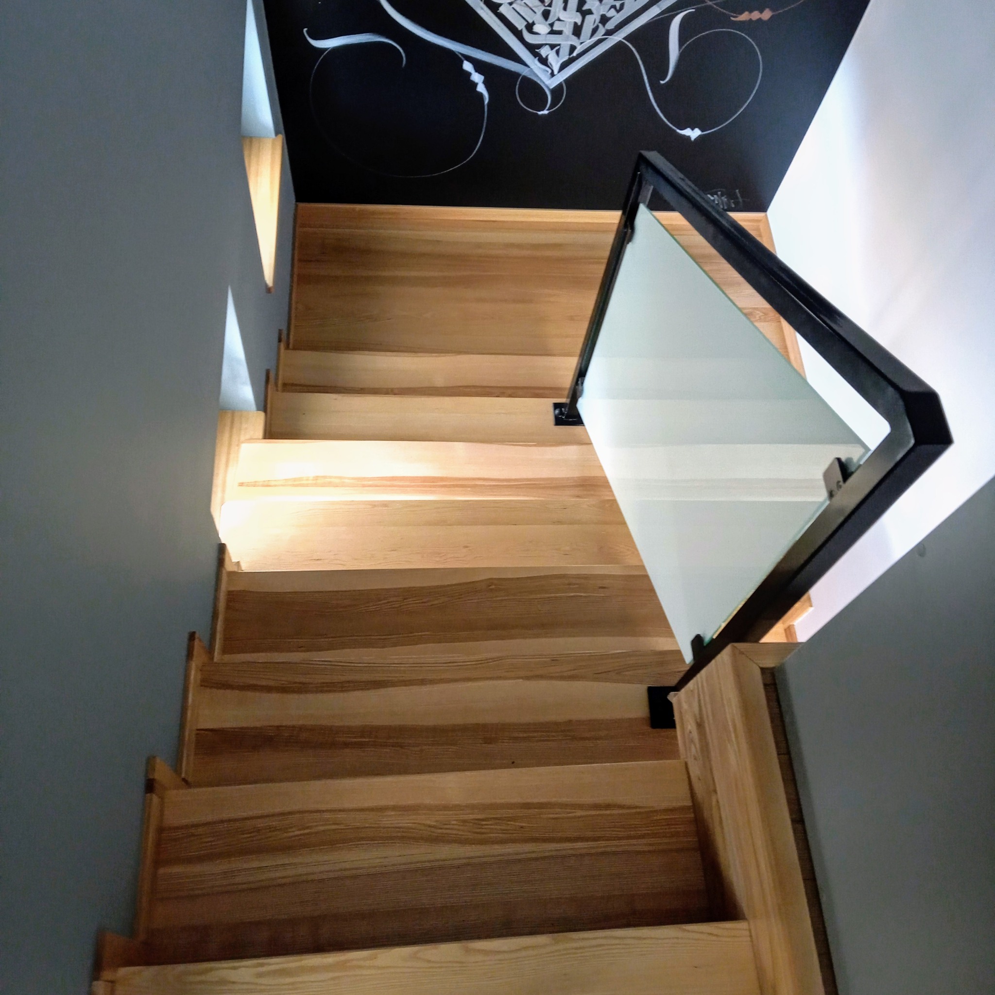 Nowoczesne schody z jasnego drewna z czarną balustradą i szklanym wypełnieniem, widok z góry, z elementami dekoracyjnymi na ścianie w tle.