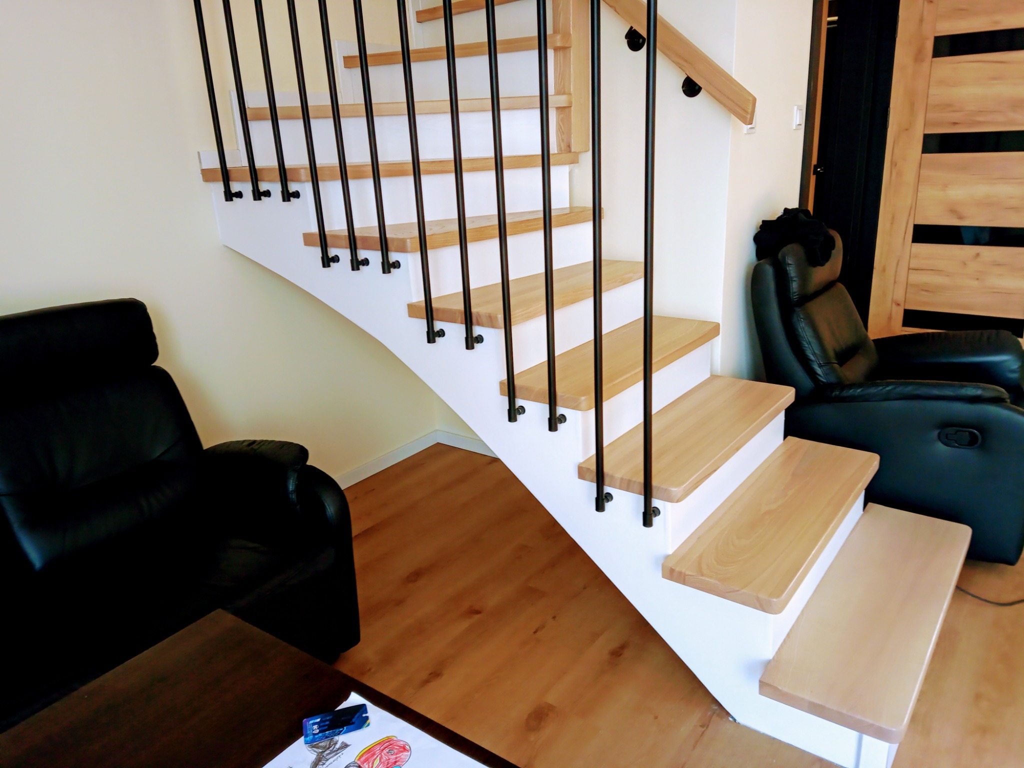 Jasne schody z drewnianymi stopniami i czarnymi balustradami w nowoczesnym wnętrzu. W tle widoczne drzwi z drewnianymi panelami i czarny fotel.