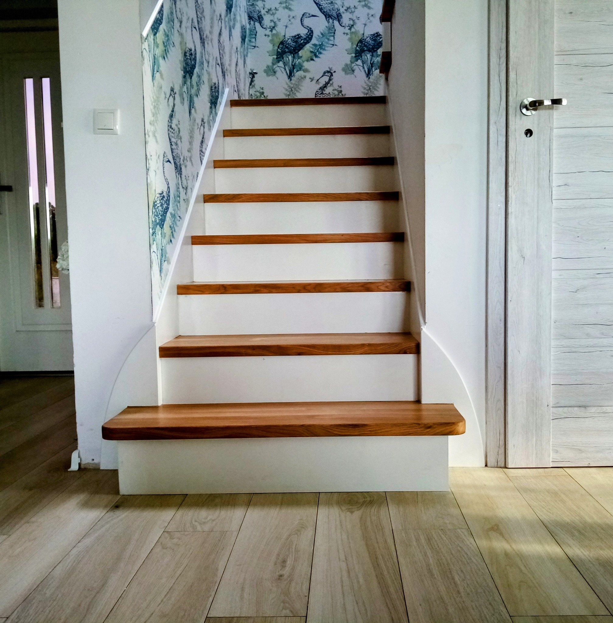 Białe schody z drewnianymi stopniami, wzorzysta tapeta z motywem ptaków, drzwi z drewnopodobną okleiną, parkiet. Ujęcie od dołu.
