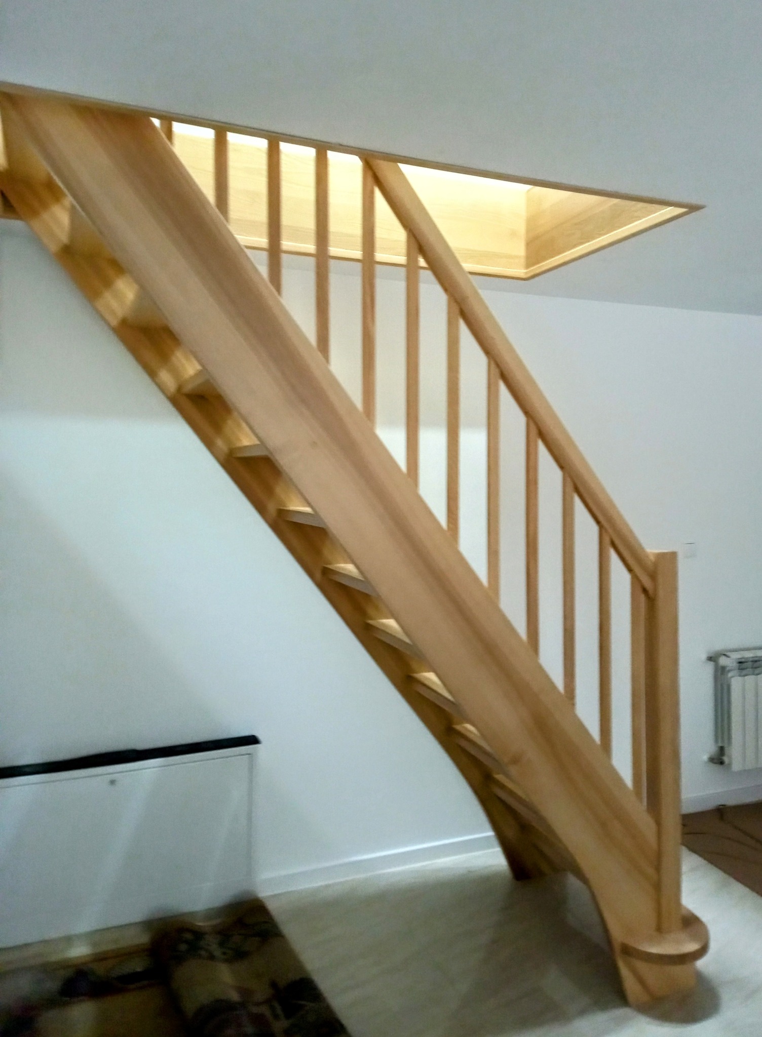 Jasne, drewniane schody z balustradą w nowoczesnym wnętrzu. Widoczny fragment podłogi i ściany pomalowanej na biało. Charakterystyczne oświetlenie w suficie.