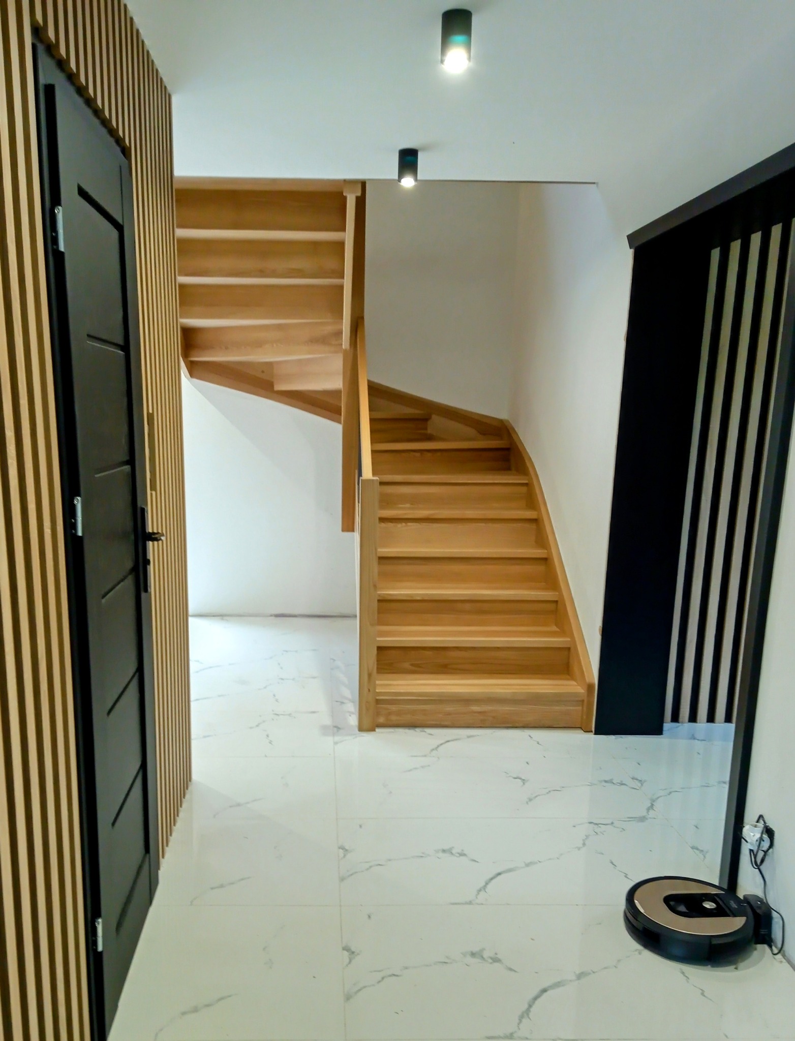 Jasne, drewniane schody z zakrętem w nowoczesnym wnętrzu z marmurową podłogą i elementami dekoracyjnymi w kolorze czarnym i drewnopodobnym.