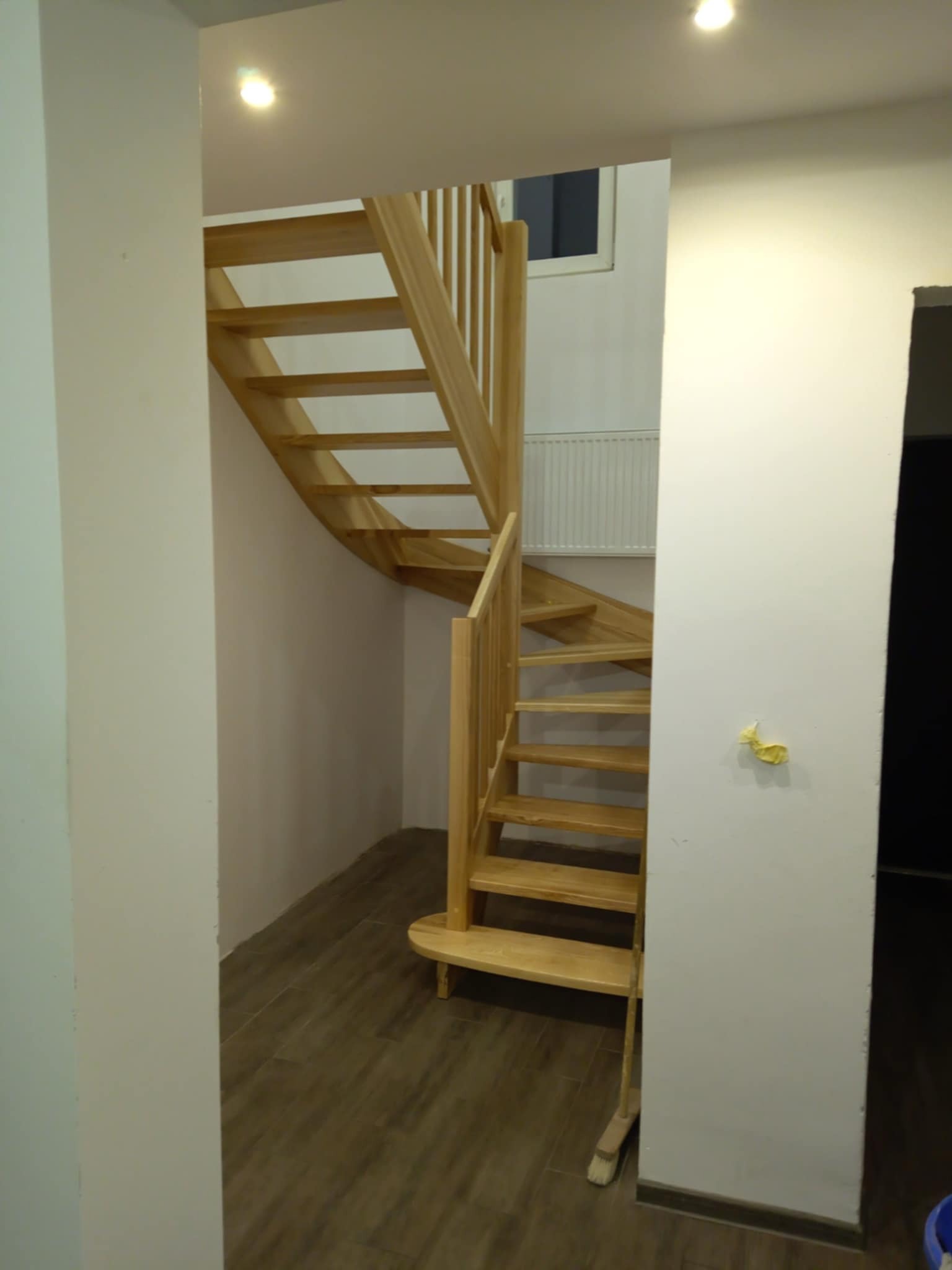 Spiralne schody drewniane z jasnego drewna, prowadzące w górę, widoczne z perspektywy dolnej części schodów. Na ścianie widoczne ślady po pracach montażowych.