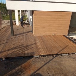 Waldek tobiaski - Drewniany taras przy nowoczesnym domu w trakcie budowy; widoczne deski, podkonstrukcja i fragment elewacji. Osoba zamiata taras.