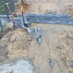 Budowa murku z szarych bloczków betonowych. Widoczny wykop, narzędzia: kielnia, poziomica, szpachelka i szlifierka kątowa. Prace ziemne w toku, częściowo ukończony fragment muru.