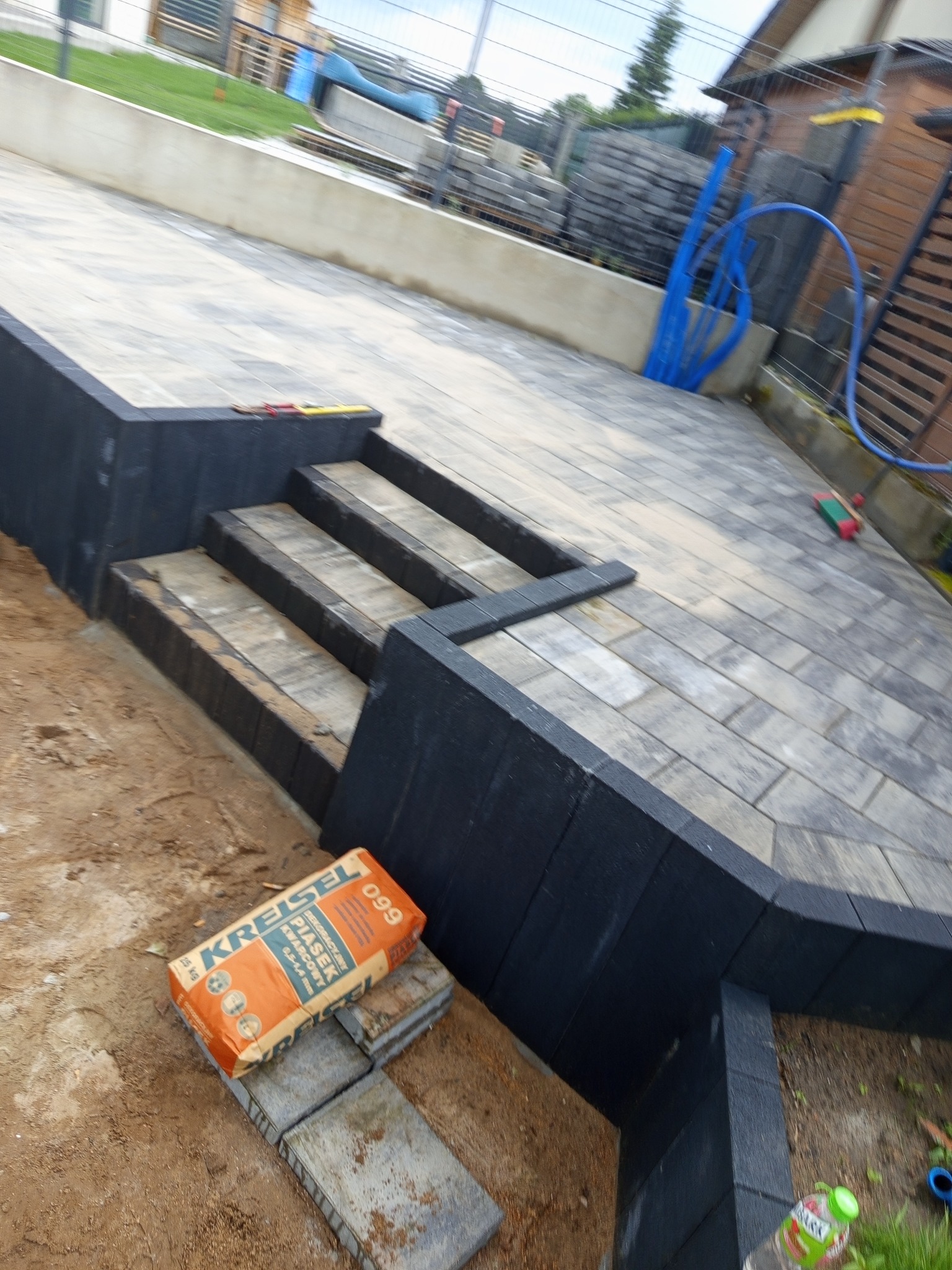 Ułożona kostka brukowa tworząca schody i taras, z workiem piasku obok. Widoczna precyzja wykonania i dbałość o detale.