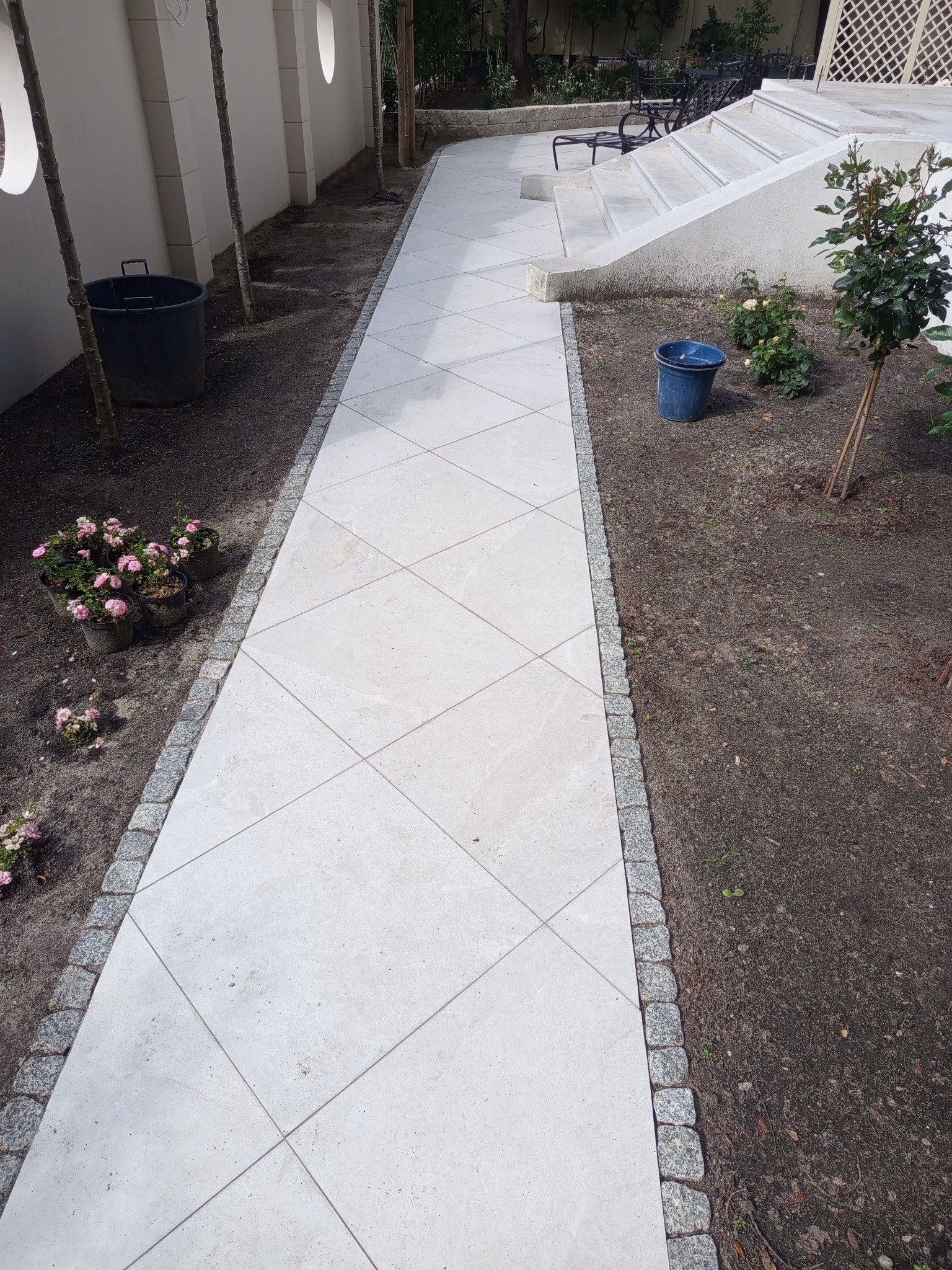 Biały chodnik z kwadratowych płyt, obramowany kostką granitową, prowadzi przez ogród z młodymi drzewkami i kwiatami doniczkowych. Schody w tle.
