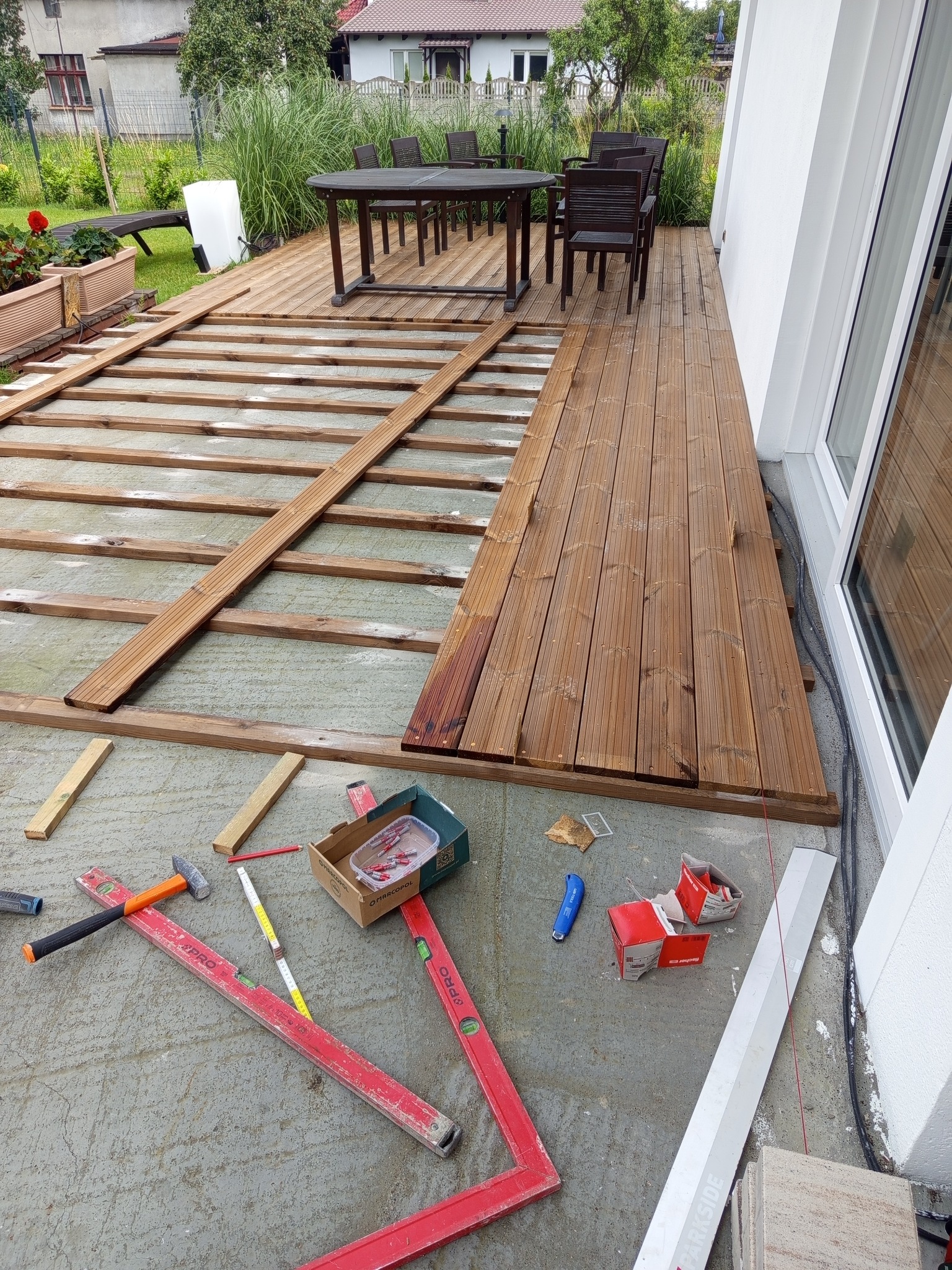 Układanie drewnianego tarasu przy domu. Częściowo ułożone deski, widoczne narzędzia pracy: poziomice, młotek, miarka. W tle stół i krzesła na gotowym fragmencie tarasu.