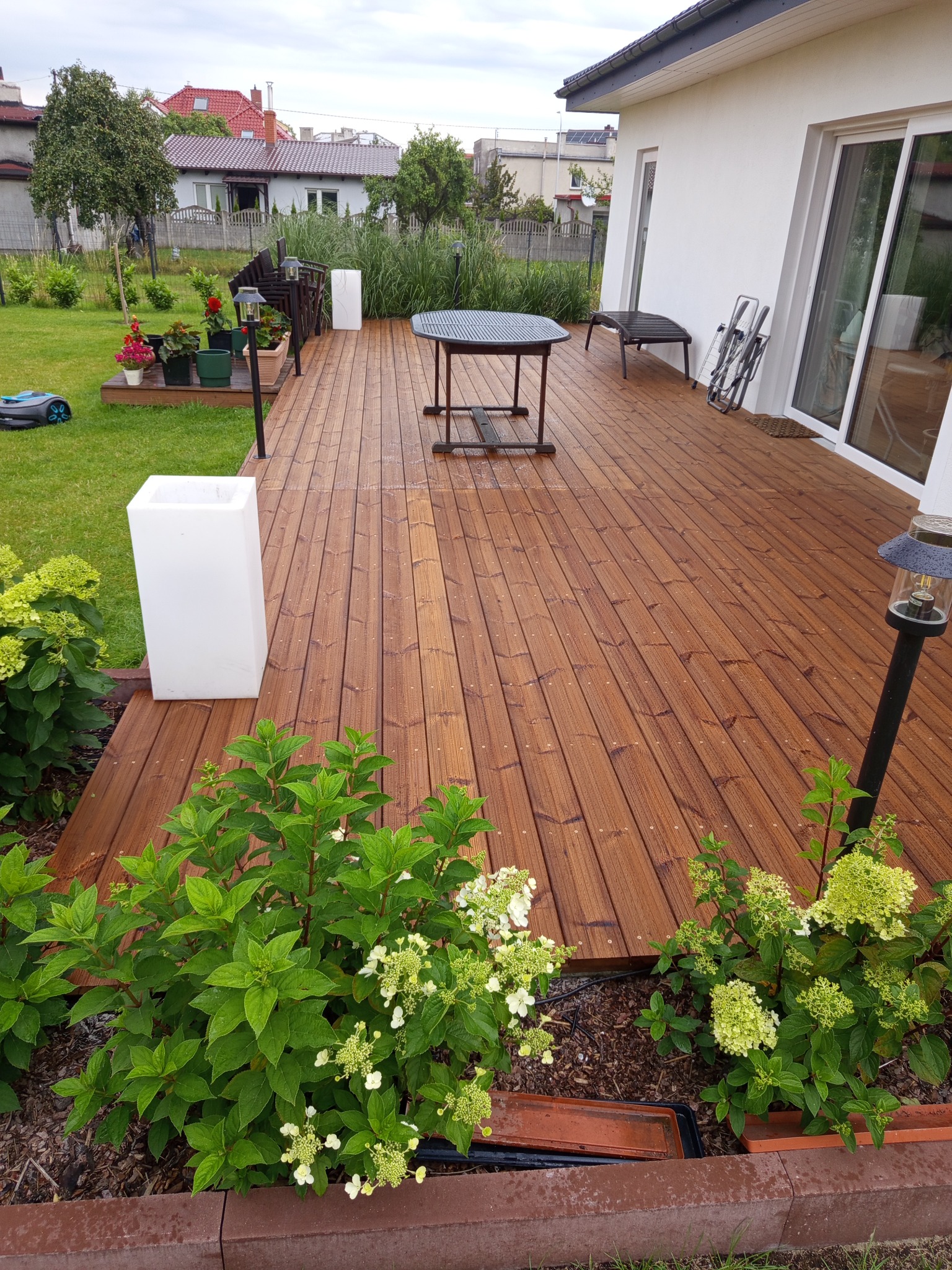 Drewniany taras przy białym domu jednorodzinnym, z roślinami wzdłuż krawędzi, stolikiem i dekoracjami. W tle trawnik z robotem koszącym.