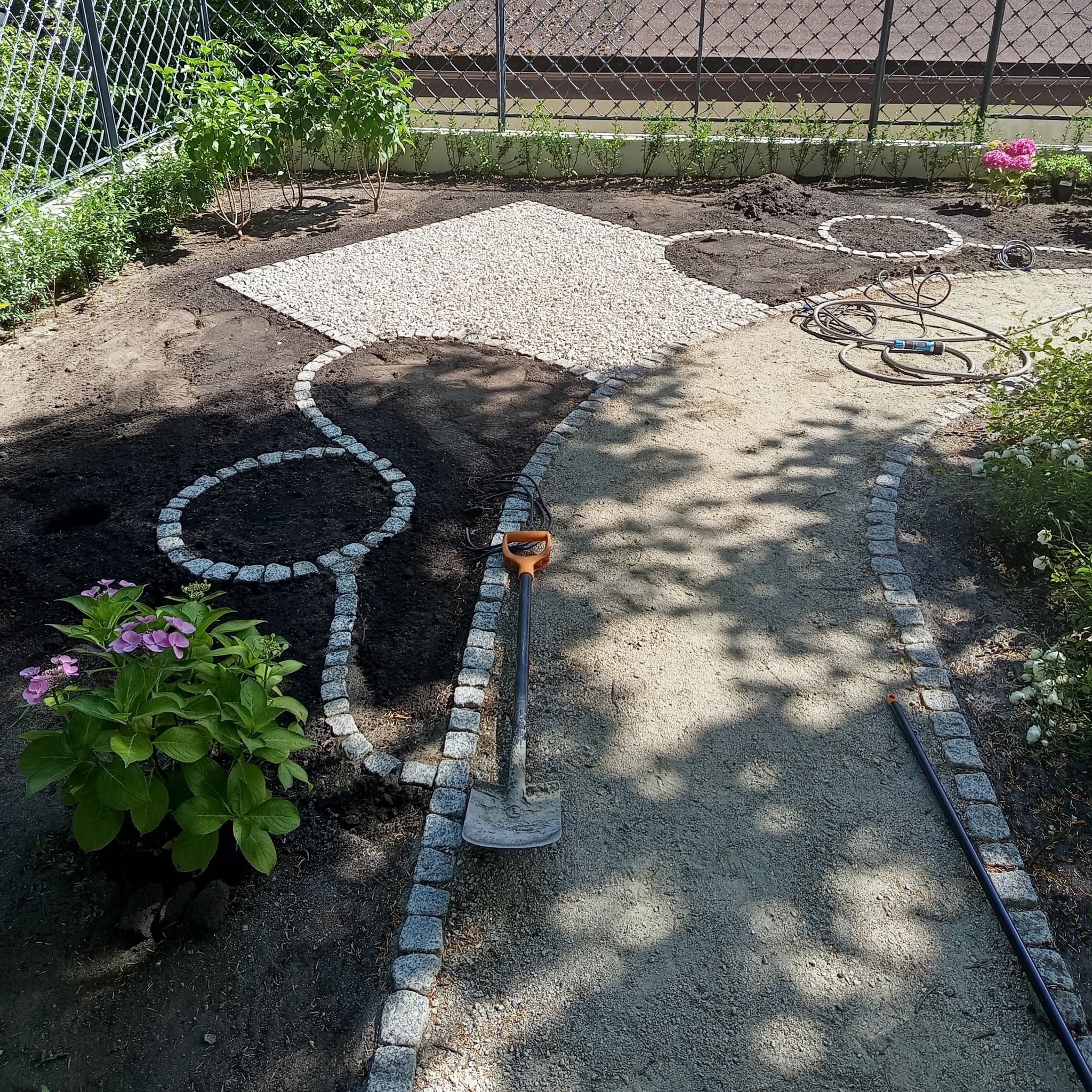 Aranżacja ogrodu w trakcie realizacji: ścieżka z kostki granitowej, rabata z białym żwirem i kwiatami. Szpadel wbity w ziemię. Widoczne elementy nawadniania.