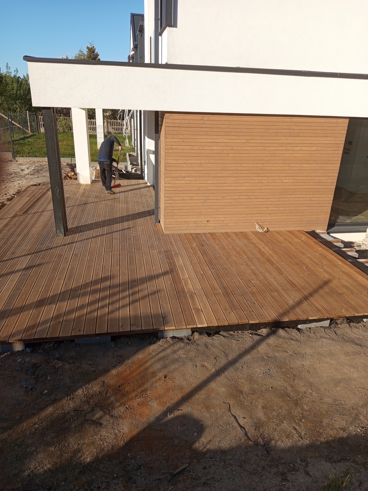 Drewniany taras przy nowoczesnym domu w trakcie budowy; widoczne deski, podkonstrukcja i fragment elewacji. Osoba zamiata taras.
