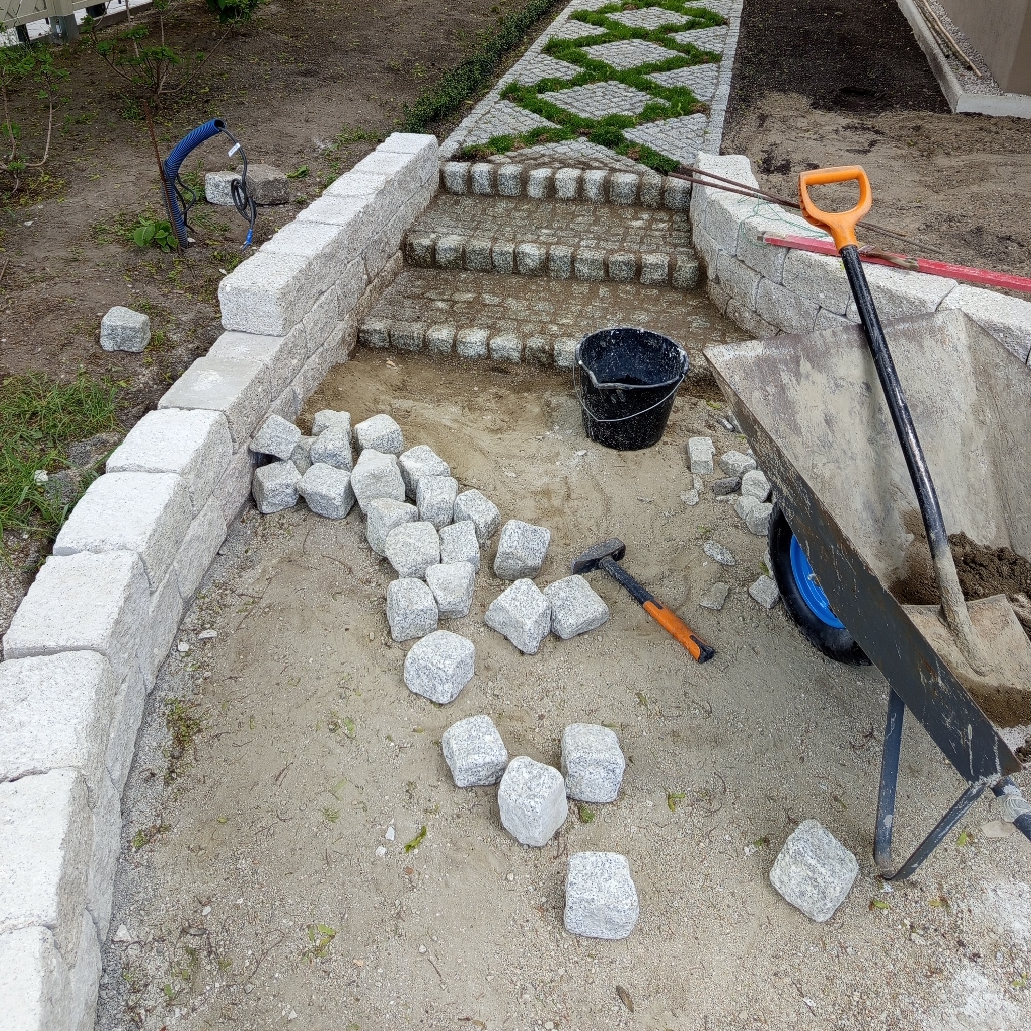 Układanie ścieżki z kostki granitowej: etap prac z rozłożonymi kostkami, widoczny fragment schodów i narzędzia (łopata, taczka, młotek).