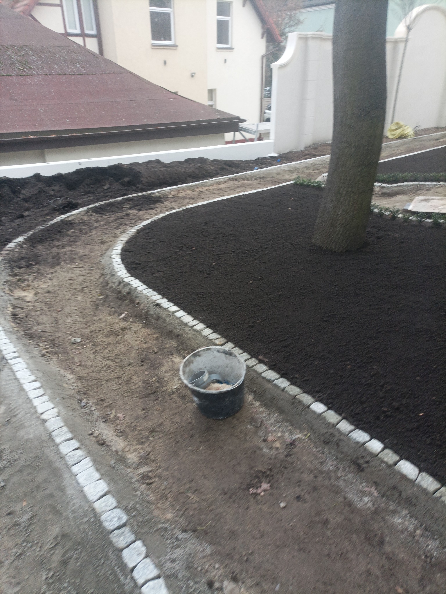 Ścieżka w ogrodzie, obrzeża z kostki granitowej, świeżo nasypana ziemia, widoczne narzędzia, etap prac ziemnych przy zakładaniu ogrodu.