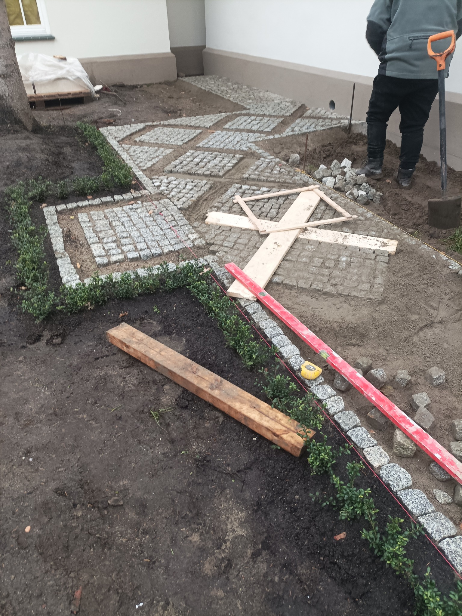 Układanie kostki granitowej w geometryczny wzór, obrzeża z bukszpanu i kostki. Widoczne narzędzia: poziomica, szpadel, drewniane stemple. Prace w toku.
