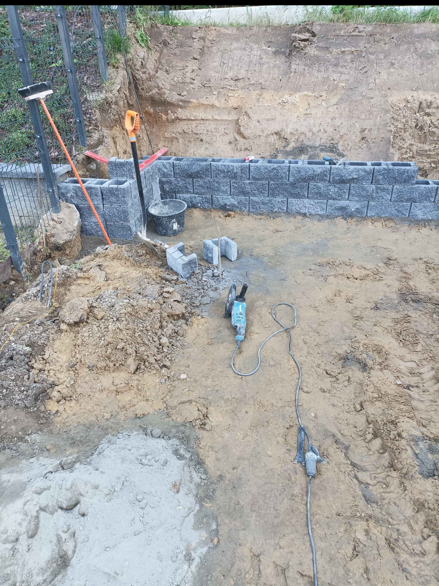 Budowa murku z szarych bloczków betonowych. Widoczny wykop, narzędzia: kielnia, poziomica, szpachelka i szlifierka kątowa. Prace ziemne w toku, częściowo ukończony fragment muru.