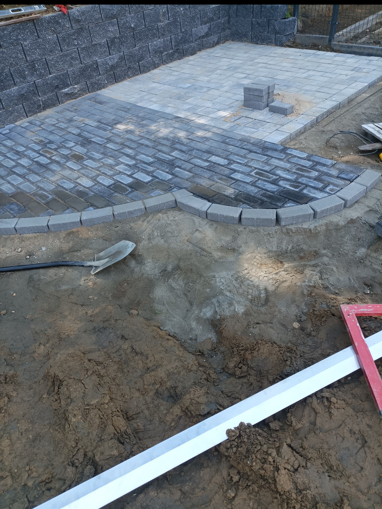 Układanie kostki: fragment nowej nawierzchni z szarej kostki brukowej o nieregularnym kształcie, widoczna łopata i poziomica. Częściowo ułożona kostka.