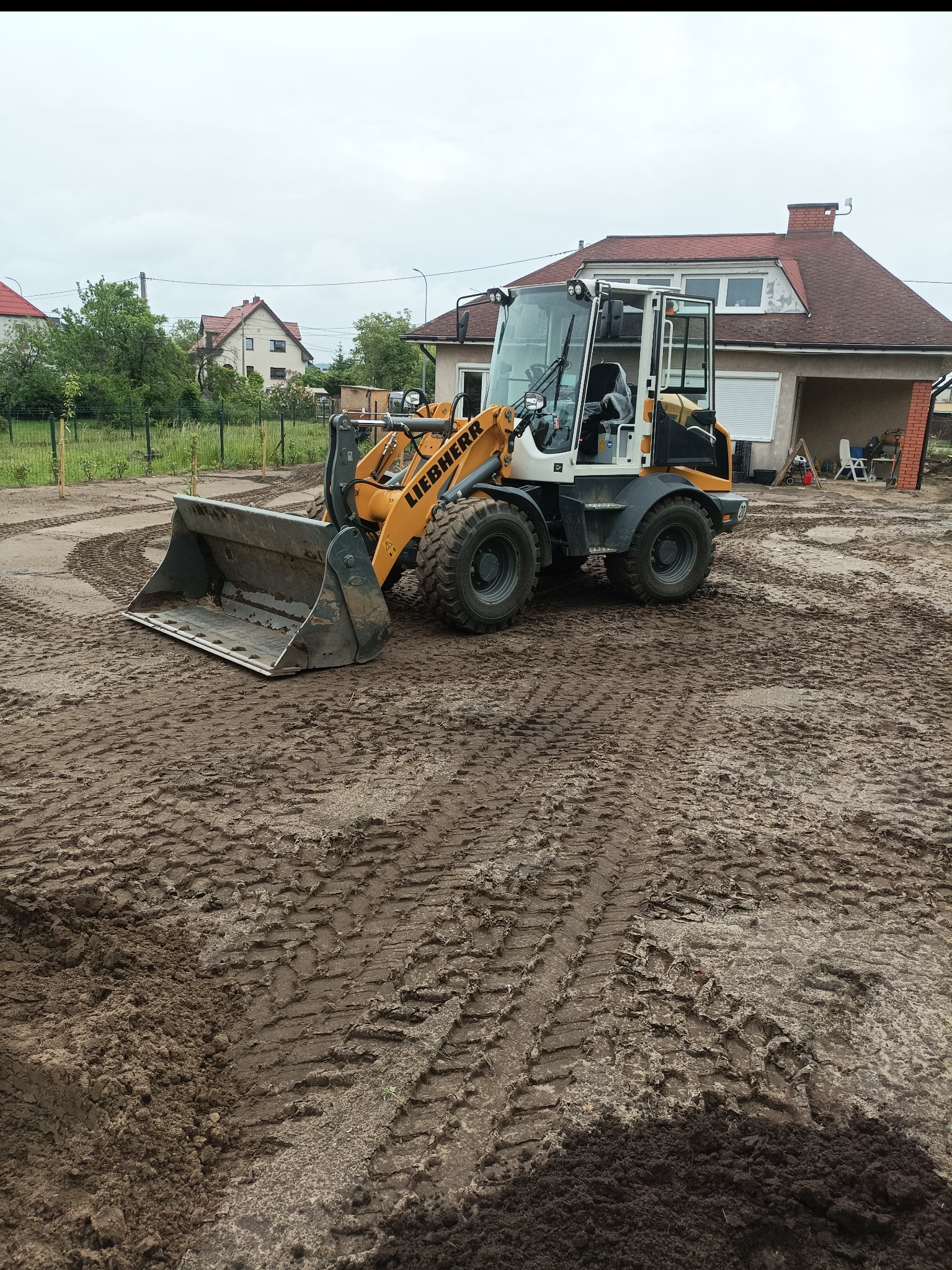 Żółta ładowarka Liebherr na placu budowy, ślady opon na ubitej ziemi, dom z czerwoną dachówką w tle. Przygotowanie terenu pod inwestycję.