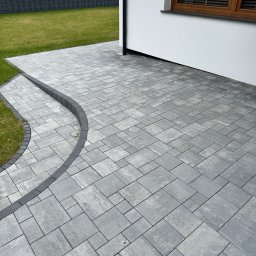 Tombud - Ułożona szara kostka brukowa na podwórku z obrzeżami, widok z góry. Nowoczesne wykończenie posesji z trawnikiem i fragmentem elewacji budynku.