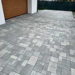 Tombud - Ułożona szara kostka brukowa na podjeździe, z widoczną bramą garażową z drewna i fragmentem zielonego ogrodzenia w tle. Różne rozmiary kostek tworzą wzór.