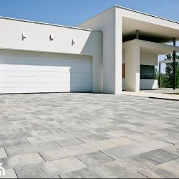 Tombud - Nowoczesny podjazd z szarej kostki brukowej przed białym domem z garażem i minimalistycznym wejściem, w słoneczny dzień. Widok z niskiej perspektywy.