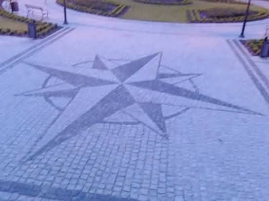 Granitowa kostka brukowa ułożona w wzór kompasu na placu, z widocznymi elementami małej architektury w tle. Nietypowy wzór i kolorystyka kostki.