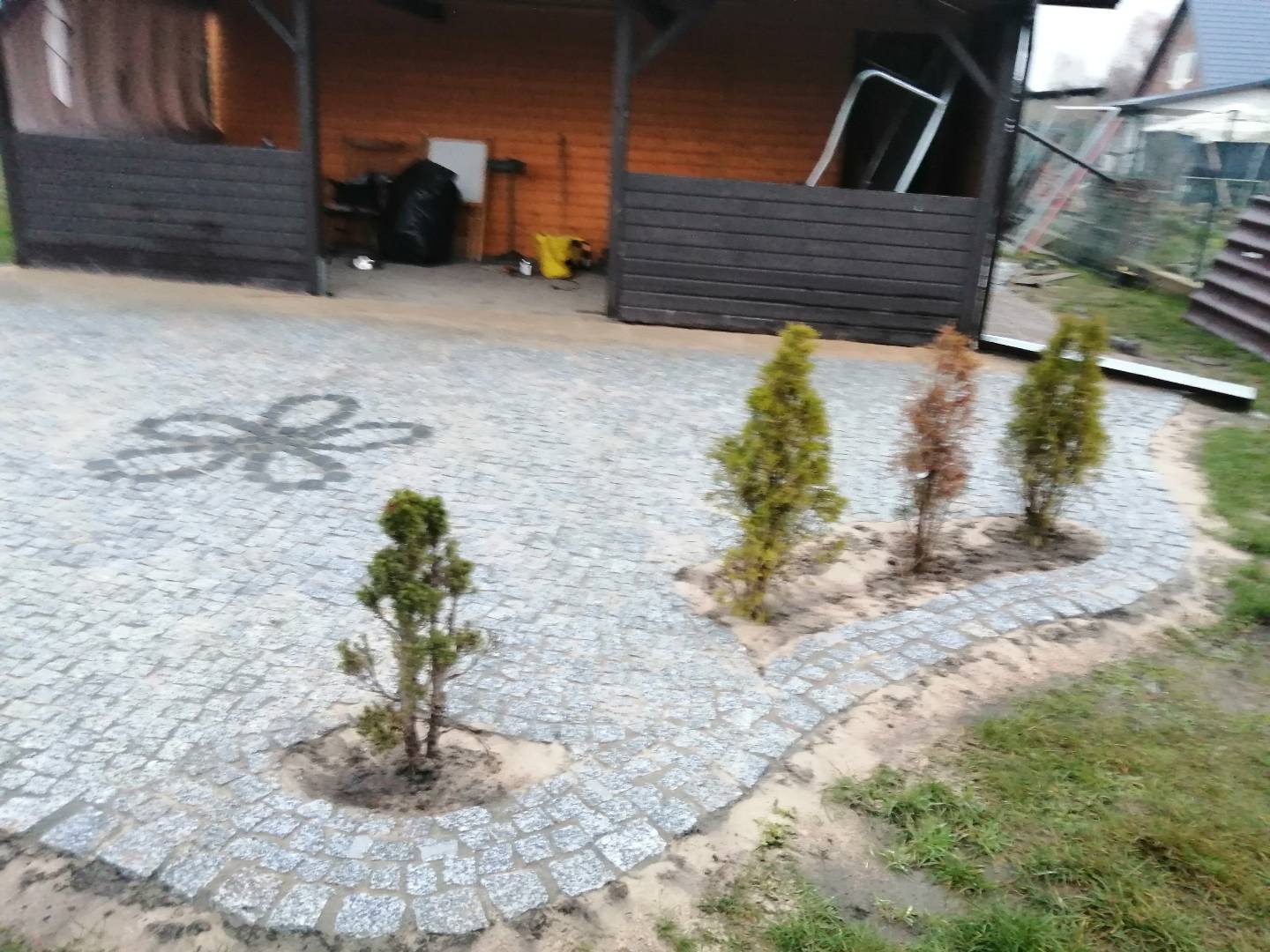 Granitowa nawierzchnia z dekoracyjnym wzorem kwiatowym i nasadzeniami iglaków, tworząca estetyczny podjazd. Widoczna altana w tle.