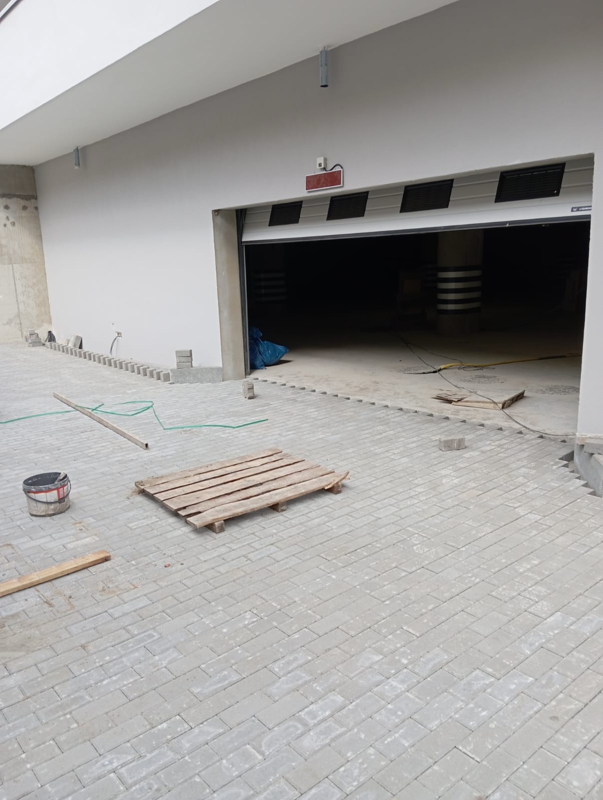 Szara kostka brukowa ułożona przed garażem, widoczna paleta, wiadro i fragmenty kostki do ułożenia. Częściowo ułożony obrzeże. Garaż z otwartą bramą.