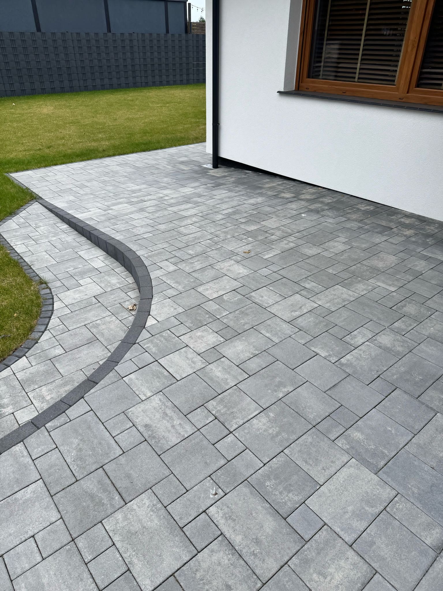 Ułożona szara kostka brukowa na podwórku z obrzeżami, widok z góry. Nowoczesne wykończenie posesji z trawnikiem i fragmentem elewacji budynku.