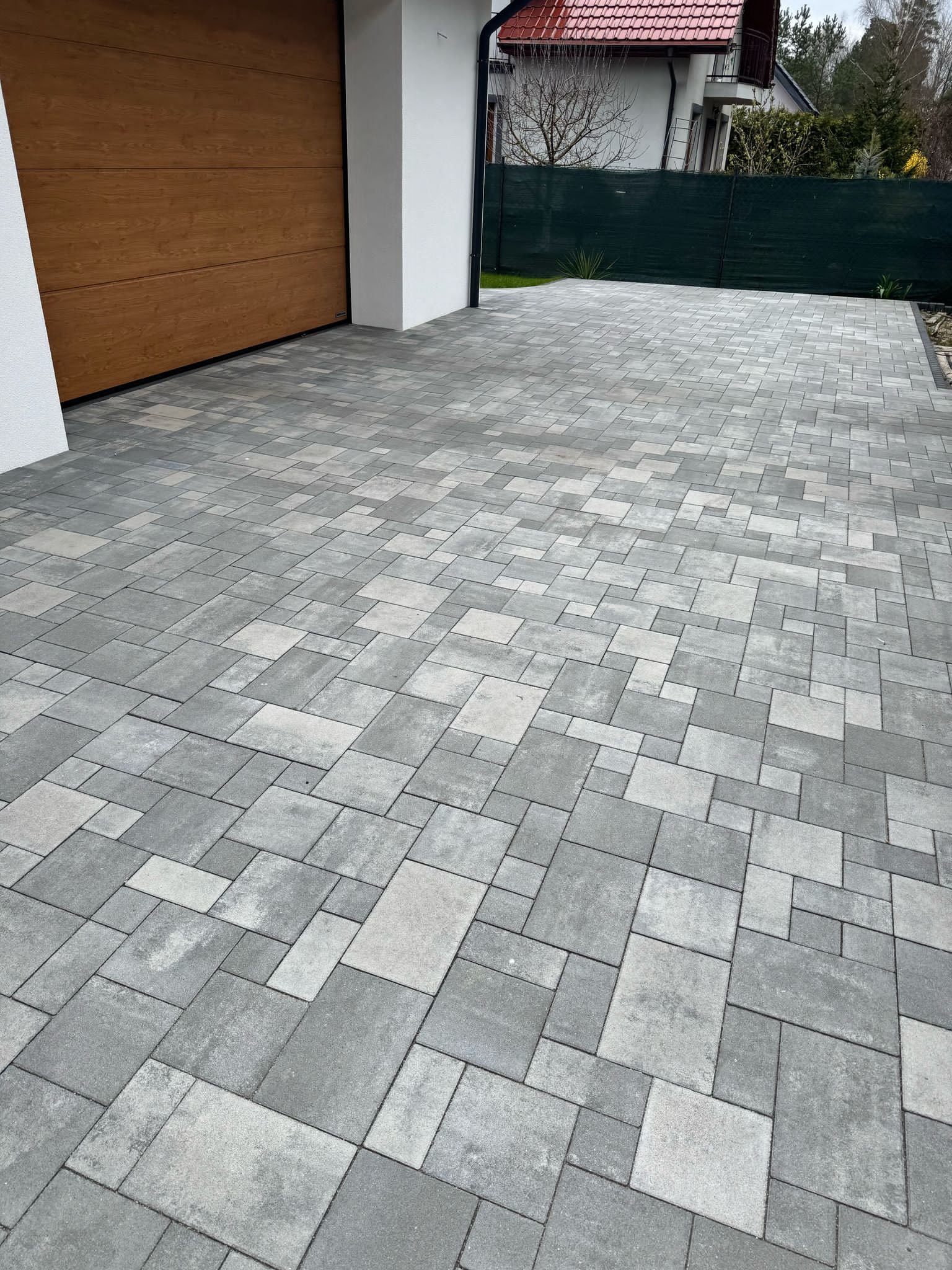 Ułożona szara kostka brukowa na podjeździe, z widoczną bramą garażową z drewna i fragmentem zielonego ogrodzenia w tle. Różne rozmiary kostek tworzą wzór.