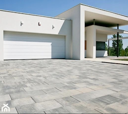 Nowoczesny podjazd z szarej kostki brukowej przed białym domem z garażem i minimalistycznym wejściem, w słoneczny dzień. Widok z niskiej perspektywy.