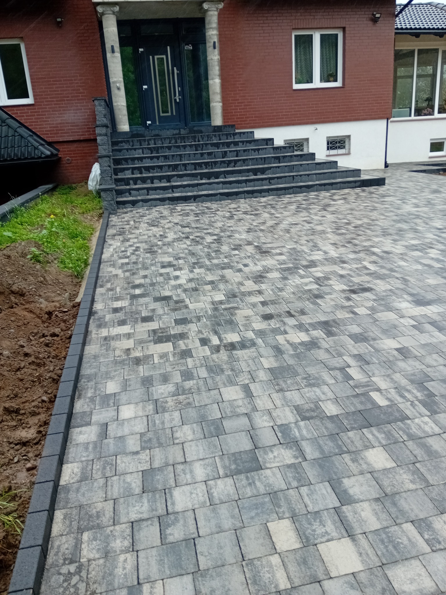 Ułożona kostka brukowa w odcieniach szarości prowadząca do schodów wejściowych domu z cegły. Widoczna krawędź trawnika i fragment elewacji budynku.