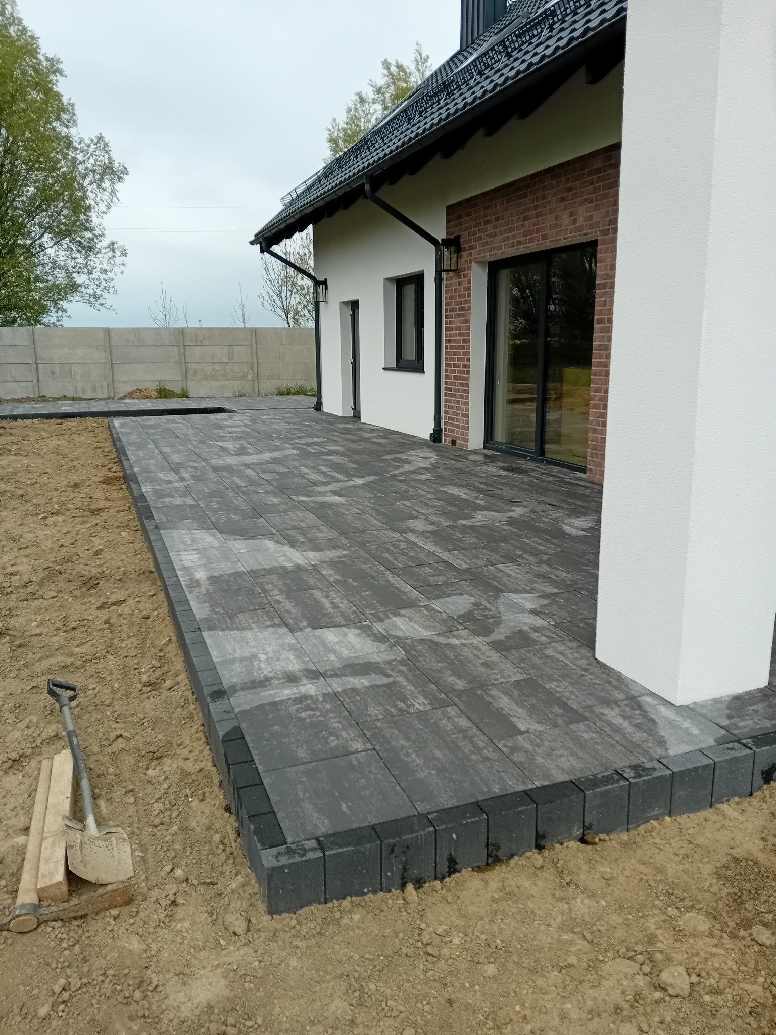 Nowoczesny taras z szarej kostki brukowej przy domu z ceglaną elewacją, widoczne narzędzia brukarskie i fragment betonowego ogrodzenia w tle.