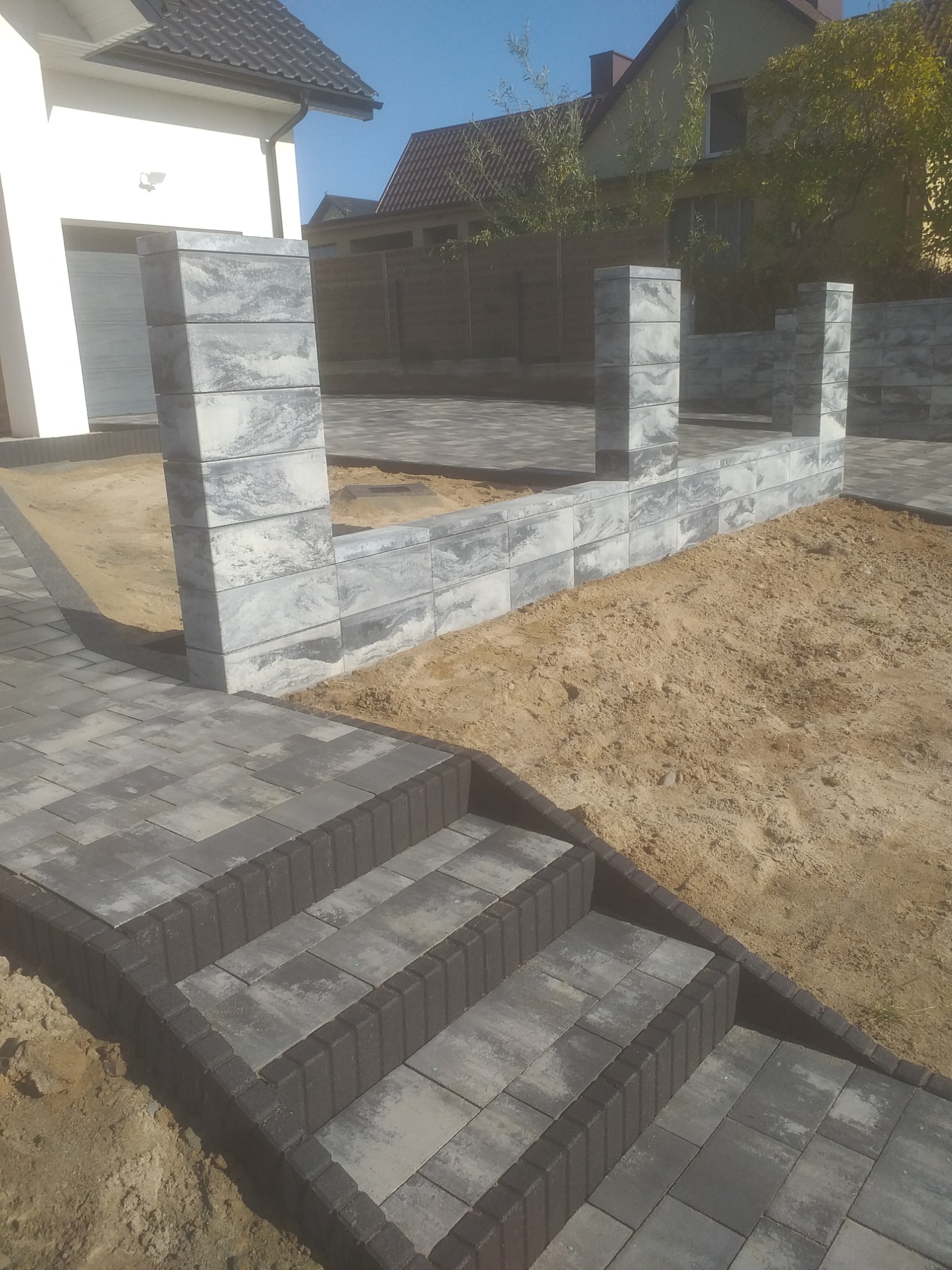 Schody i fragment ogrodzenia z szarej kostki brukowej na posesji. Widoczne elementy domu w tle. Świeżo ułożona kostka, obok piasek.