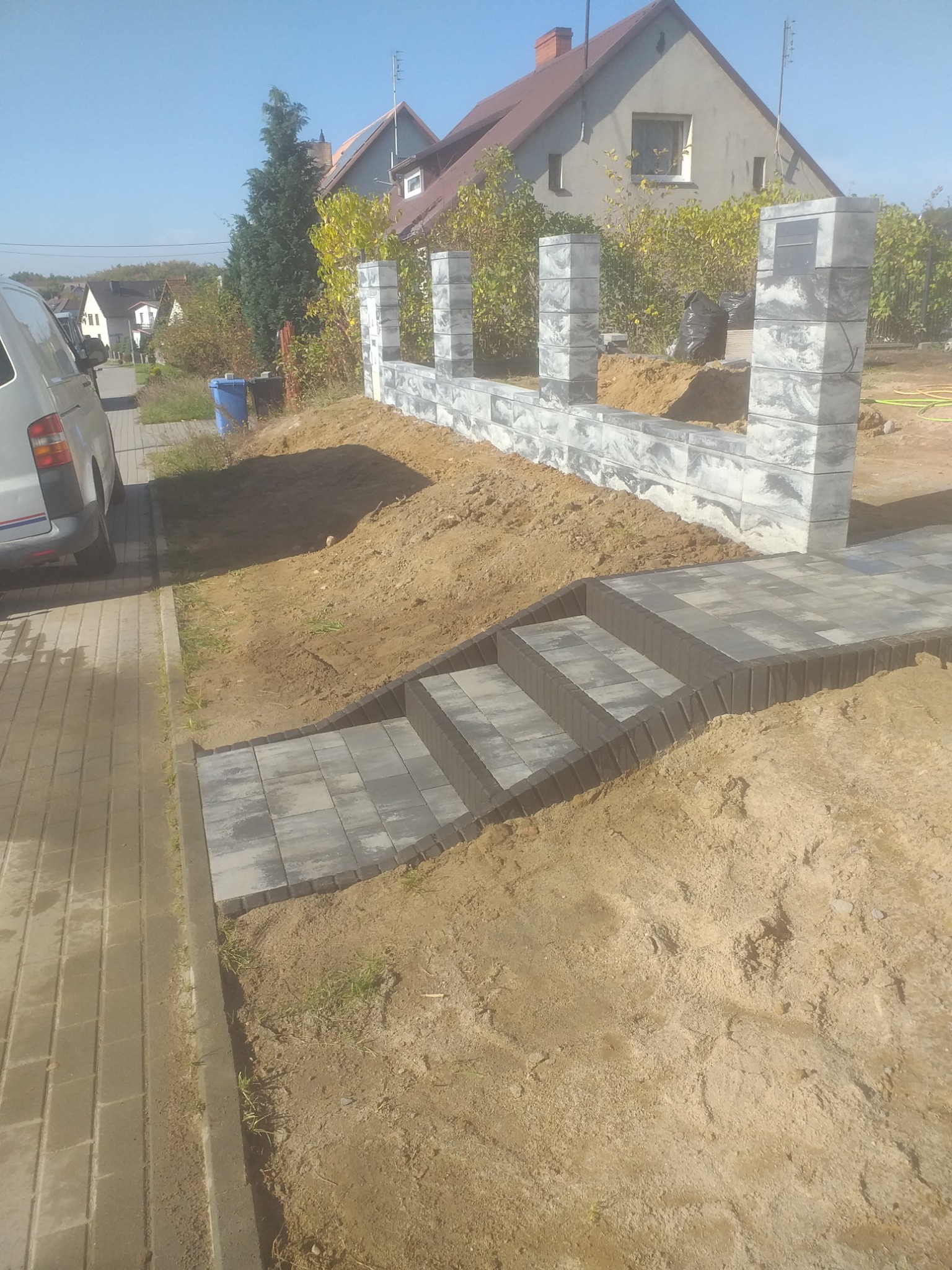 Schody z kostki brukowej prowadzące do ogrodzenia z bloczków w szarościach, widoczne prace ziemne i fragment chodnika z kostki. Słoneczny dzień.