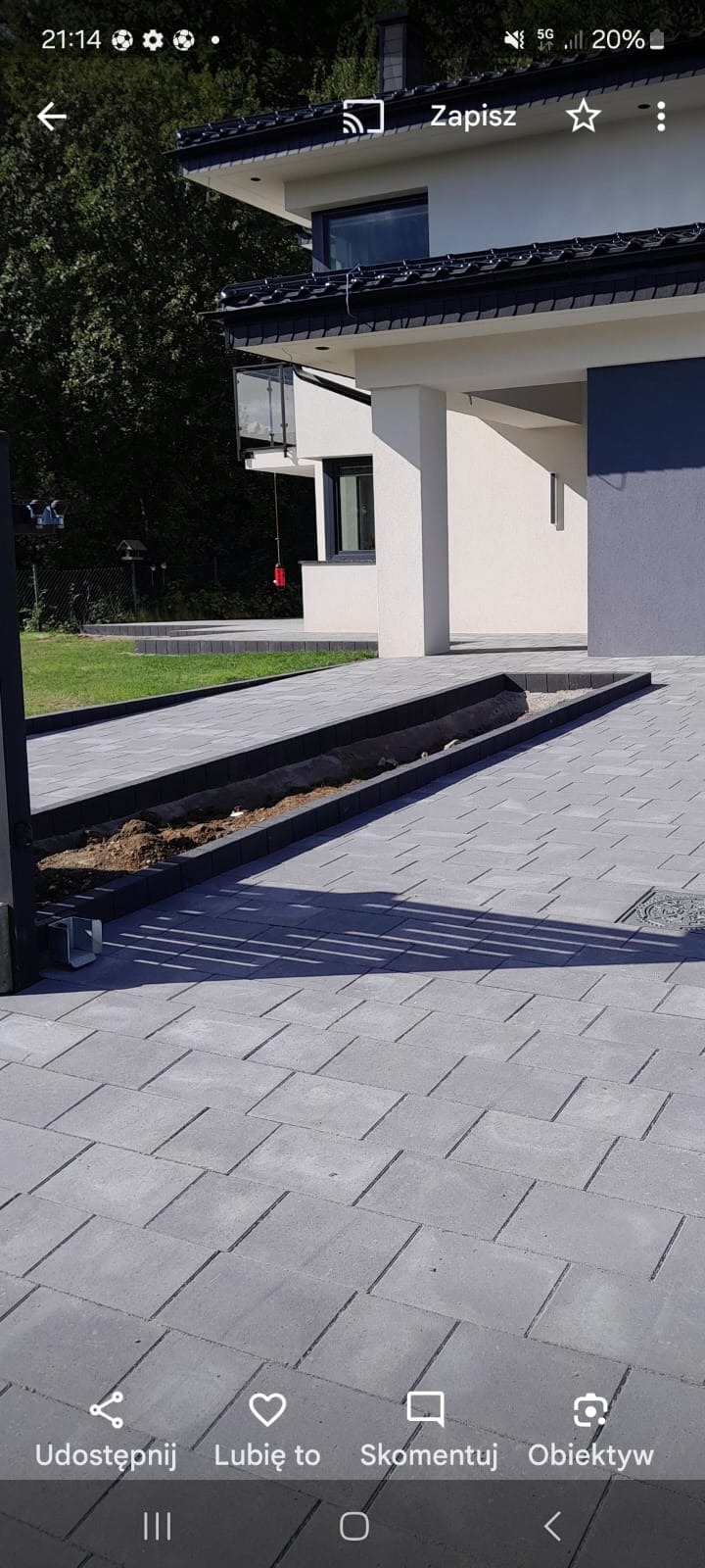 Szara kostka brukowa ułożona na podjeździe, z fragmentem wykopu pod instalację. Widoczny budynek mieszkalny w tle i cień bramy wjazdowej. Regularny wzór ułożenia kostki.