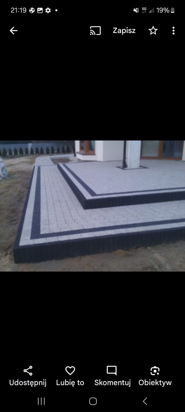 Ułożona kostka brukowa w odcieniach szarości z ciemnym obramowaniem tworząca schody i ścieżkę przy jasnym budynku. Widoczne krzewy i trawnik w tle.